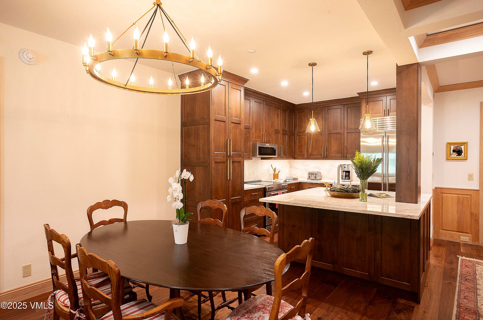 This interior shot showcases a well-appointed kitchen and dining area. The kitchen features dark wood cabinetry, stainless steel appliances, and a marble countertop island. The dining area includes a large oval table surrounded by wooden chairs, illuminated by a decorative chandelier, creating a warm and inviting atmosphere.