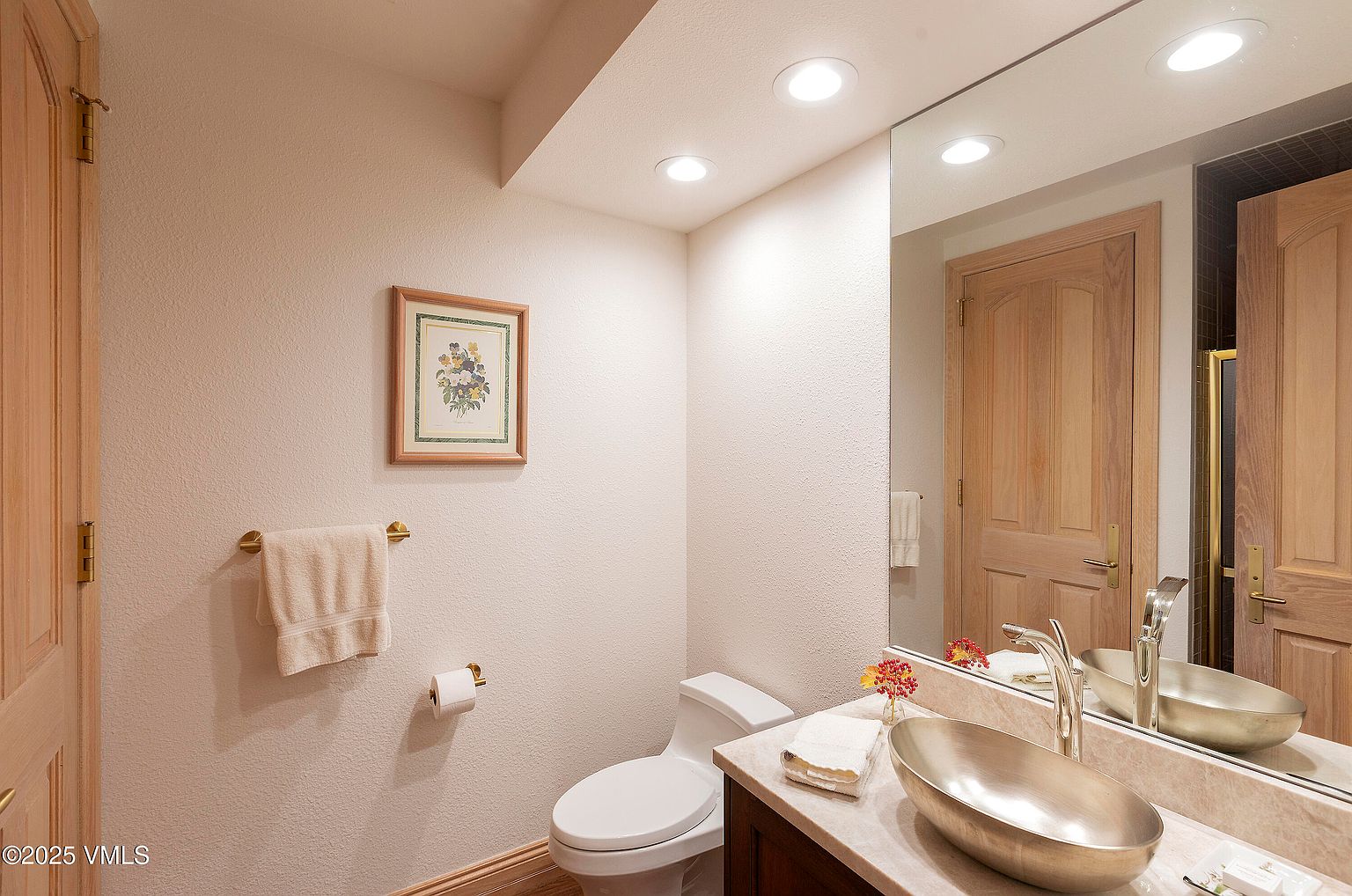 This is a well-lit guest bathroom featuring a unique oval-shaped sink, a marble countertop, and a large mirror that reflects a wooden door. The walls are painted in a neutral tone, complemented by a framed floral picture and a towel rack with a neatly folded towel. The overall impression is clean, functional, and subtly elegant.