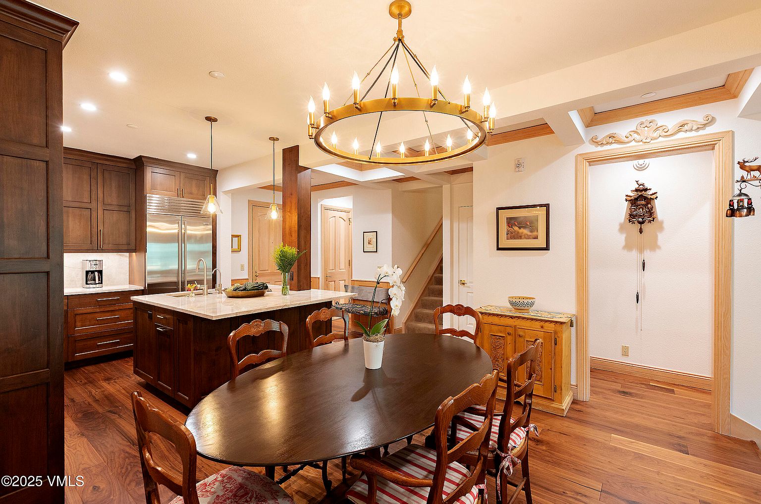 This is an interior shot of a dining room featuring a large oval wooden table surrounded by wooden chairs with patterned cushions. A striking chandelier hangs above the table, providing warm lighting. The room also includes hardwood floors, a built-in cabinet, and a glimpse into an adjacent kitchen area, creating a cozy and inviting atmosphere.