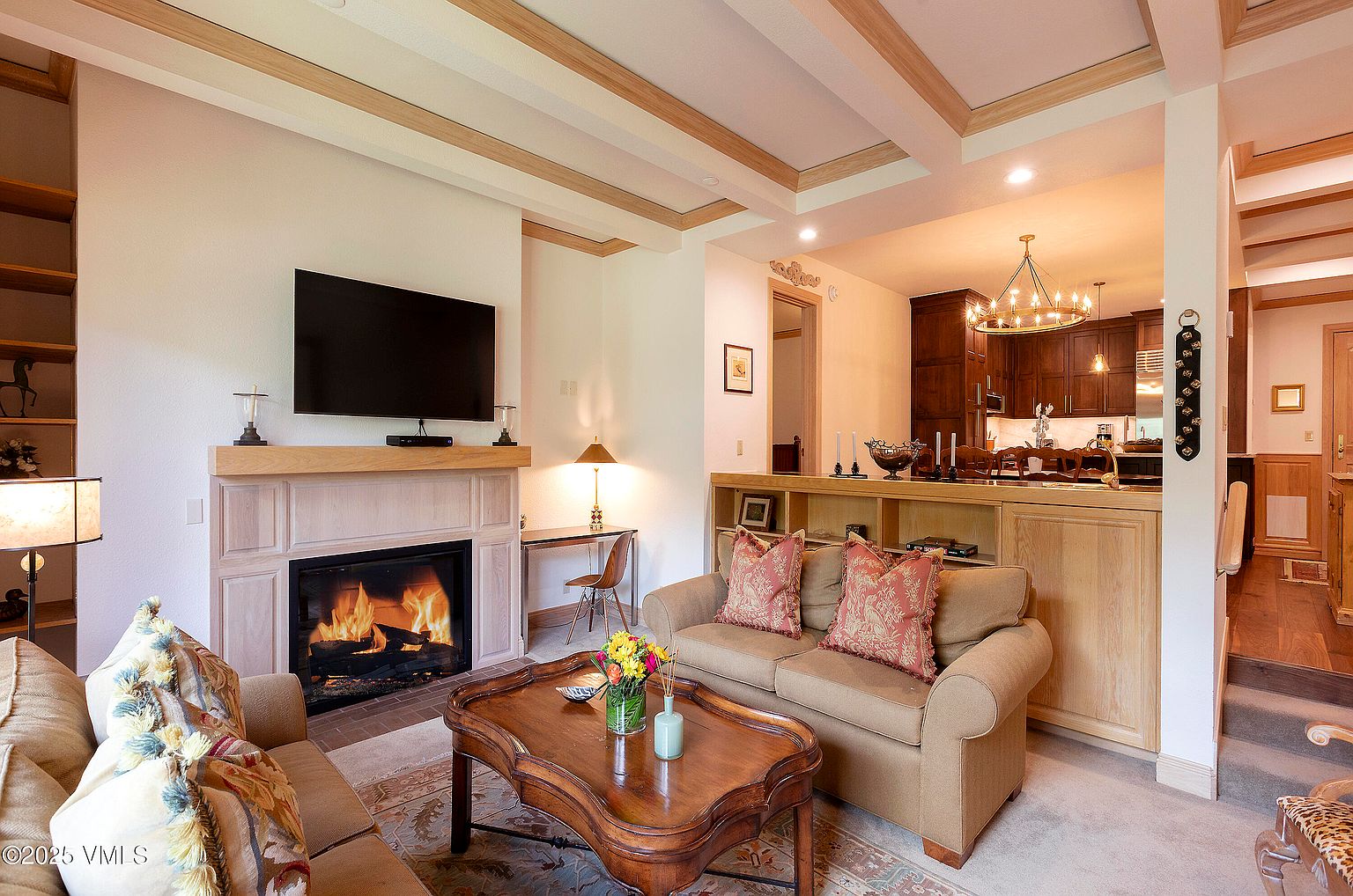 This is an interior shot of a cozy living room featuring a fireplace with a lit fire, a flat-screen TV mounted above the mantel, and comfortable seating. The room opens to a kitchen area, creating an open-concept living space. The coffered ceiling with wooden beams adds architectural interest, and the overall ambiance is warm and inviting.