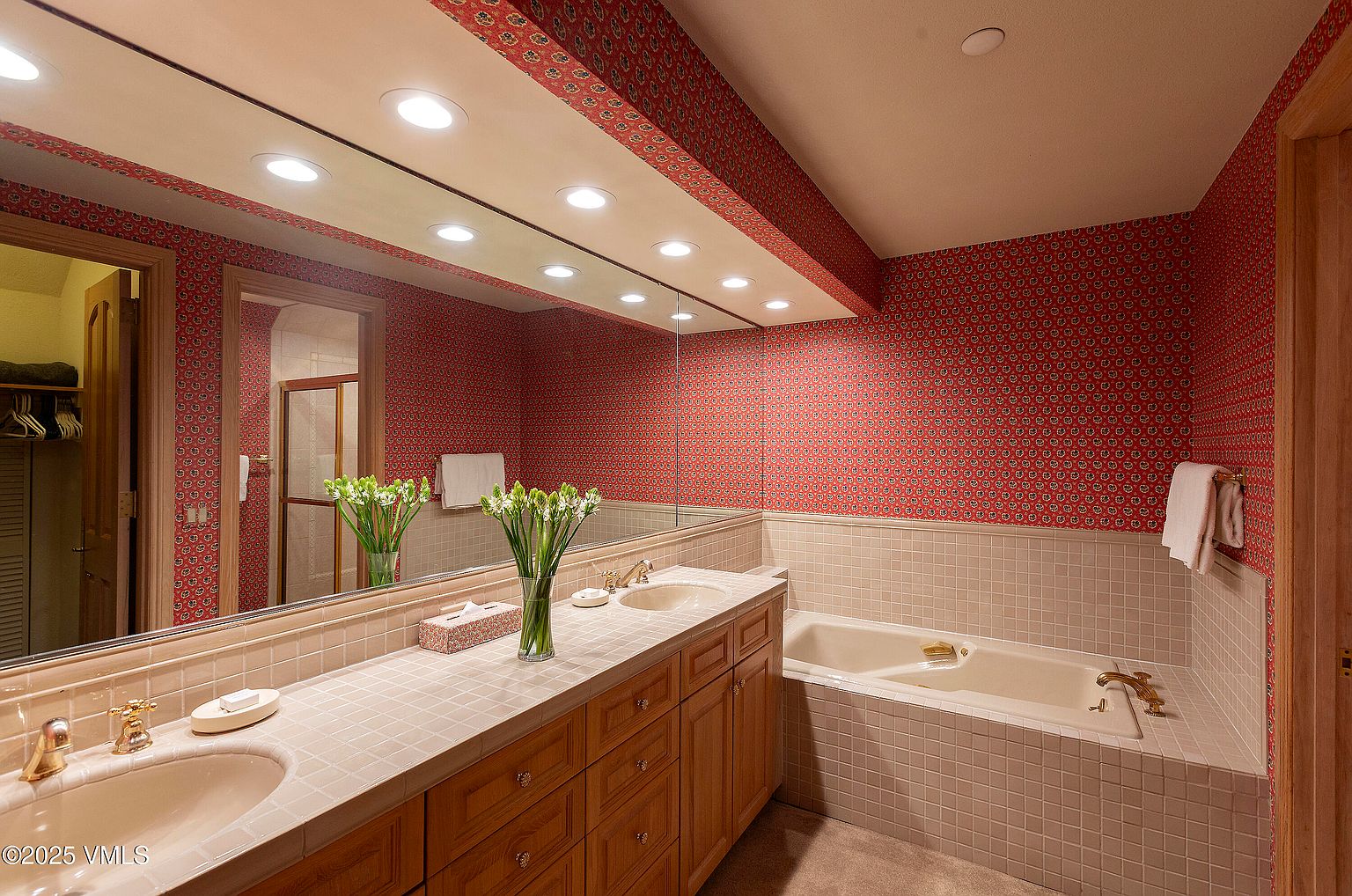 This is a primary bathroom featuring a double vanity with a tiled countertop and wooden cabinets. A large mirror spans the length of the vanity, reflecting the red patterned wallpaper that covers the walls and ceiling. A tiled bathtub is situated to the right, and a doorway is visible on the left, suggesting access to other areas of the primary suite.