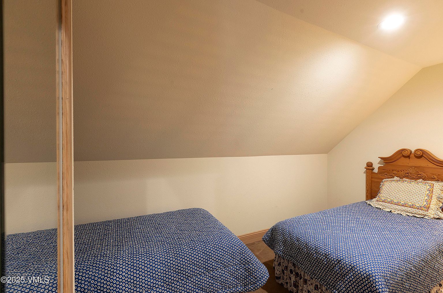 This is an interior shot of a guest bedroom featuring two twin beds with blue patterned bedding. The room has a sloped ceiling and neutral-colored walls, creating a cozy and simple atmosphere. One of the beds has a decorative wooden headboard, adding a touch of traditional style to the space.