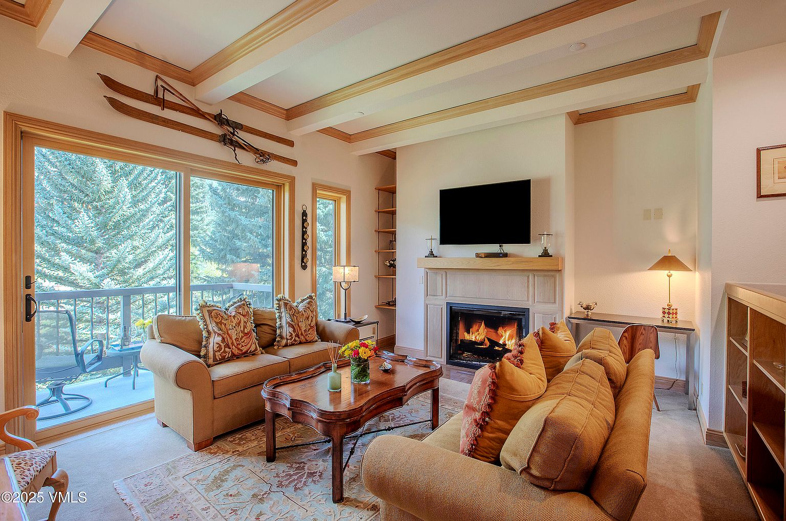 This is an interior shot of a cozy living room featuring two sofas, a wooden coffee table, and a fireplace with a mounted television above it. The room has a warm and inviting atmosphere, enhanced by the natural light coming through a large sliding glass door that leads to a balcony. The decor includes decorative pillows, a patterned rug, and wooden ceiling beams, adding to the room's charm and character.