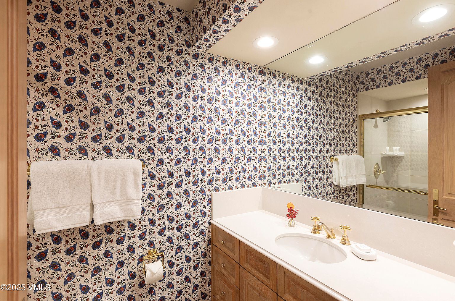 This is a bathroom featuring a vanity with wood cabinets and a white countertop, a white sink, and gold-toned fixtures. The walls are covered in a patterned wallpaper with blue and red floral motifs. A large mirror reflects the vanity area and a shower with a glass door, creating a sense of spaciousness.