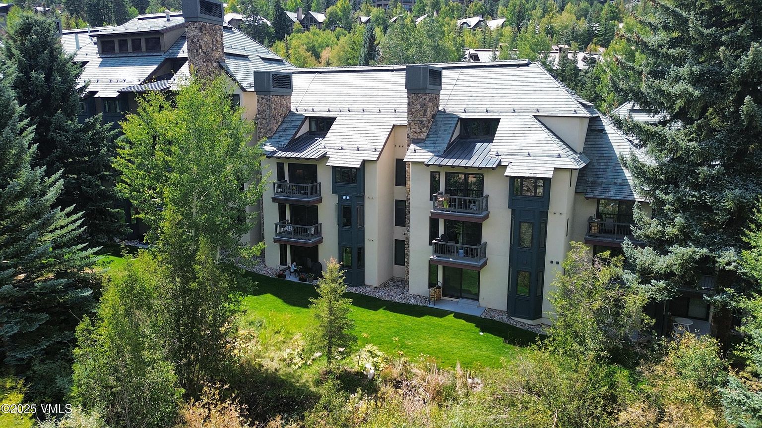 This aerial view showcases a multi-story residential building with a stone chimney and multiple balconies, surrounded by lush greenery and mature trees. The building features a neutral color palette with dark accents, complemented by a well-maintained lawn. The overall impression is one of luxury and tranquility, ideal for potential buyers seeking a serene living environment.