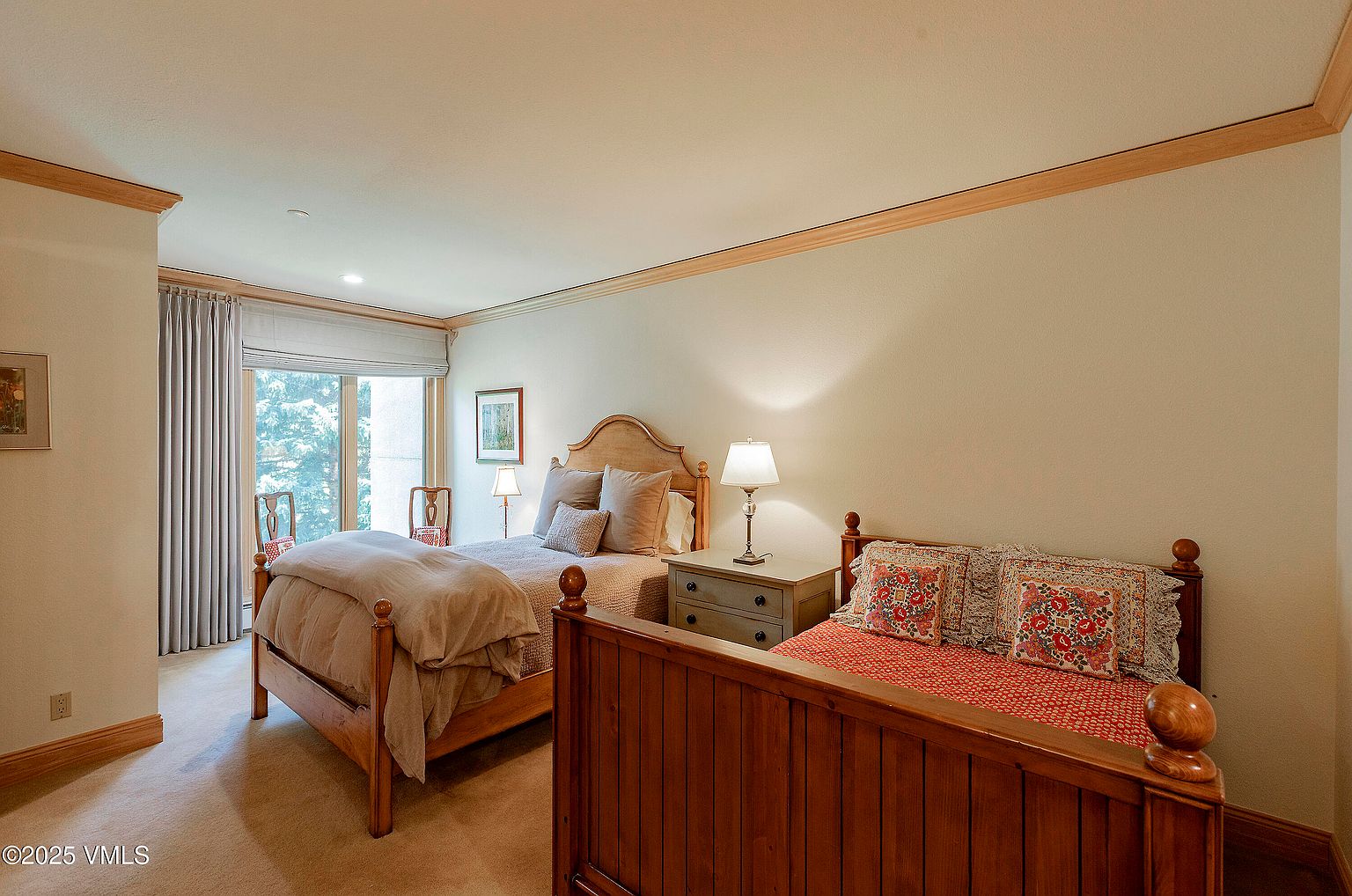 This interior shot showcases a guest bedroom featuring two beds, one full and one twin, with wooden frames and neutral-toned bedding. A bedside table with a lamp sits between the beds, and a window with curtains provides natural light. The room has a cozy and inviting atmosphere, suitable for a comfortable stay.