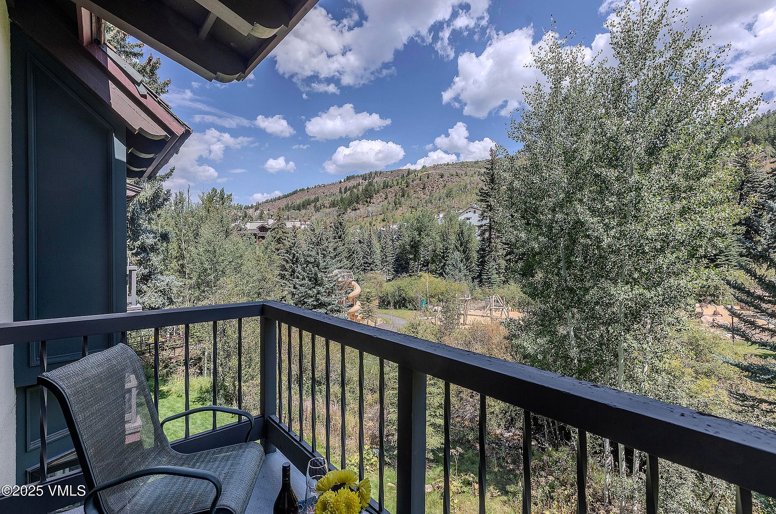 This image showcases a charming balcony view, complete with a cozy chair, a bottle of wine, and a bouquet of yellow flowers. The balcony overlooks a lush landscape with trees and a glimpse of a playground, set against a backdrop of a hill under a partly cloudy sky. The scene evokes a sense of relaxation and tranquility, highlighting the outdoor living space as a desirable feature of the property.