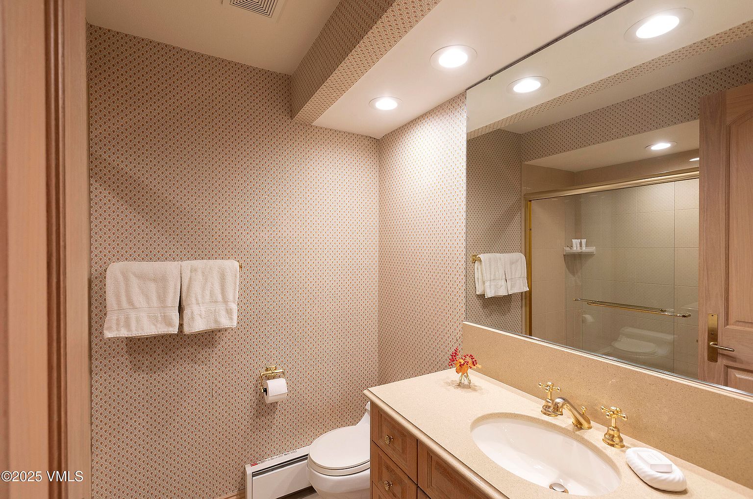 This is a bathroom featuring patterned wallpaper, a vanity with a sink, and a toilet. The vanity has a light-colored countertop and wooden cabinets. A mirror above the vanity reflects a shower with a glass door. The overall style is traditional and well-maintained.