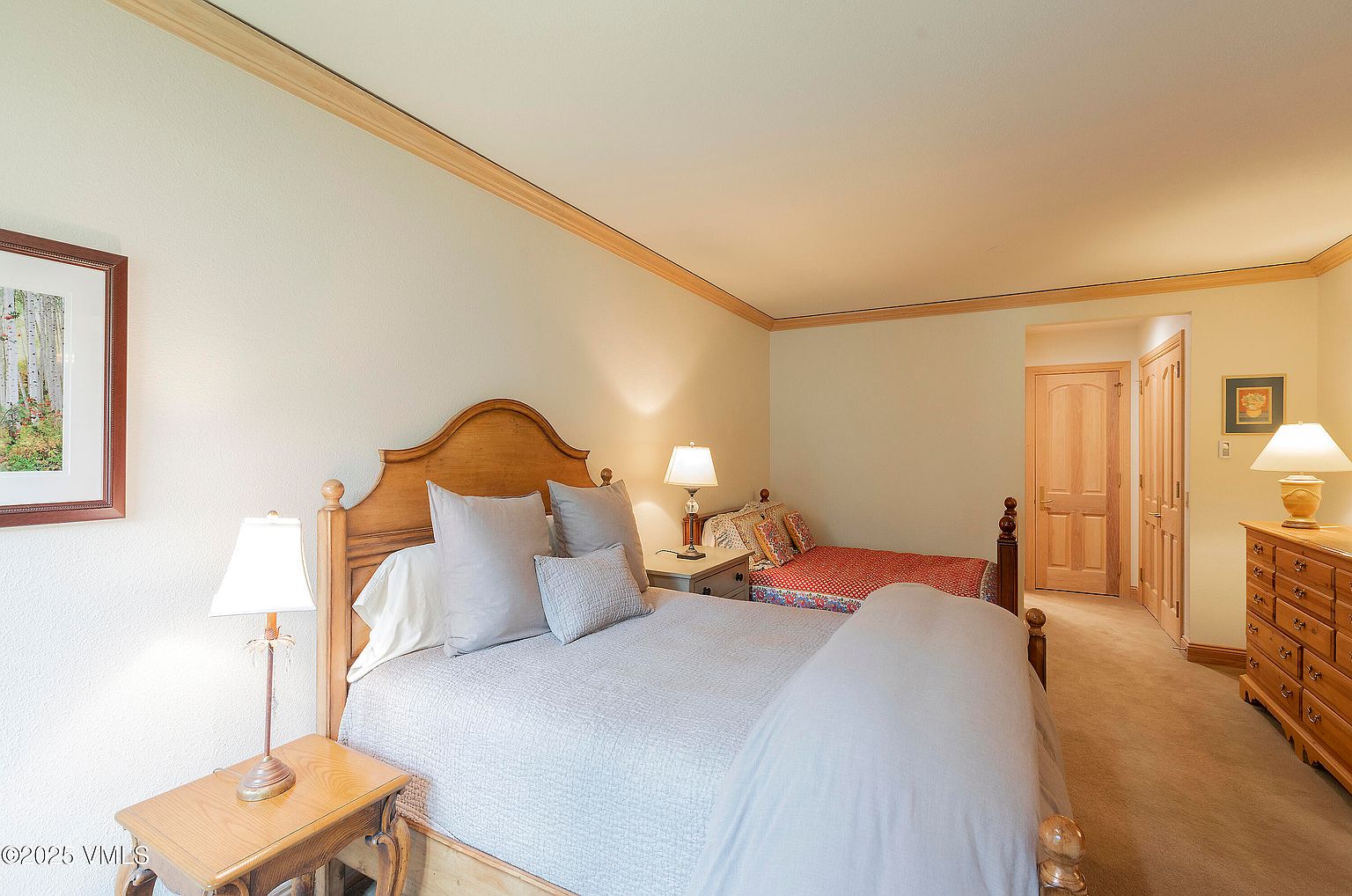 This is a cozy guest bedroom featuring two beds, one queen and one twin, with wooden frames and neutral-toned bedding. The room is illuminated by bedside lamps, and a wooden dresser is visible in the background. The walls are painted in a warm, neutral color, complemented by wooden trim, creating a comfortable and inviting atmosphere.