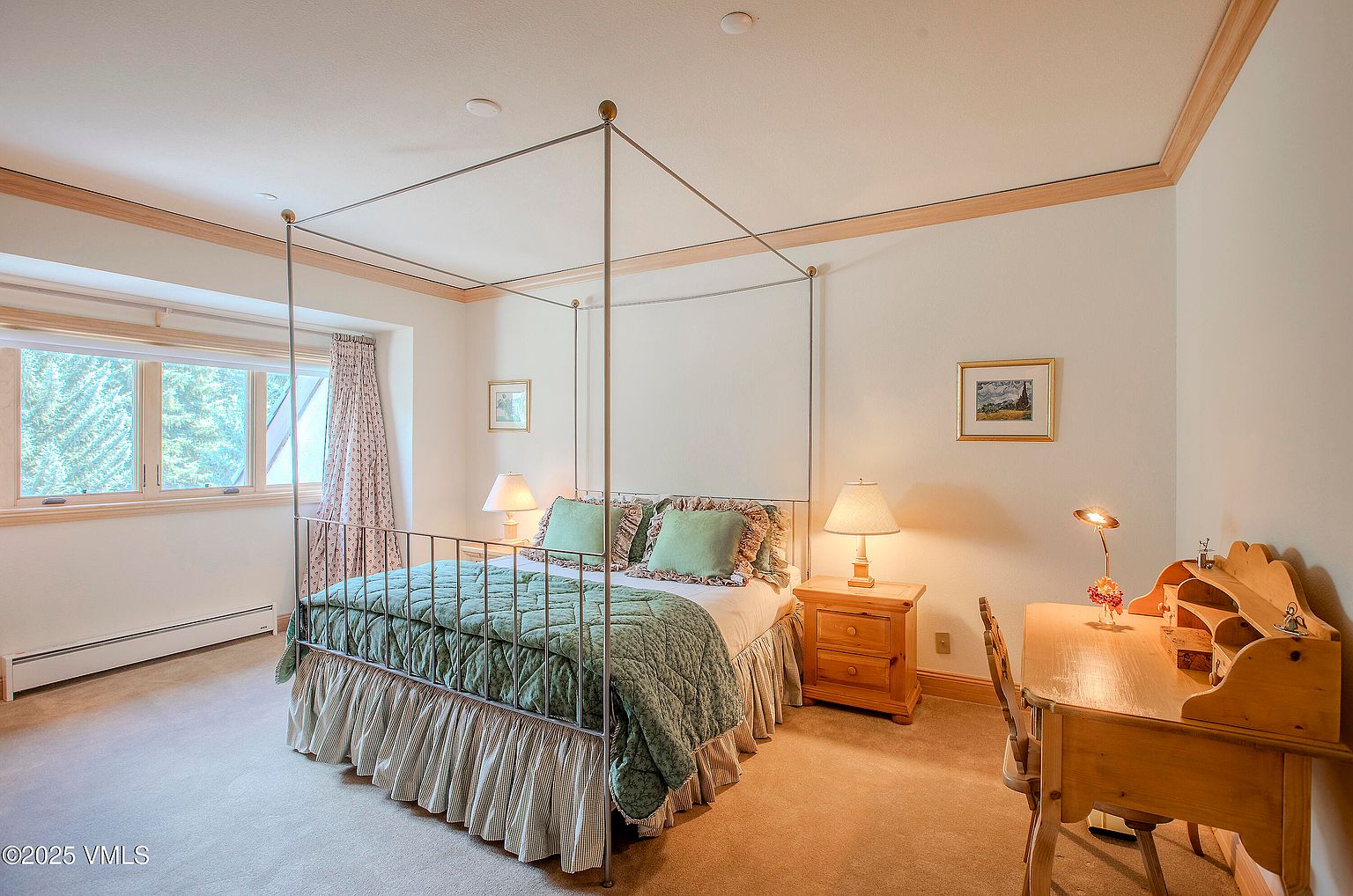 This is a cozy bedroom featuring a four-poster bed with a green quilted comforter and matching pillows. A wooden desk and chair are positioned to the right, complemented by a bedside table with a lamp. The room is well-lit, with natural light streaming through a window, creating a warm and inviting atmosphere.