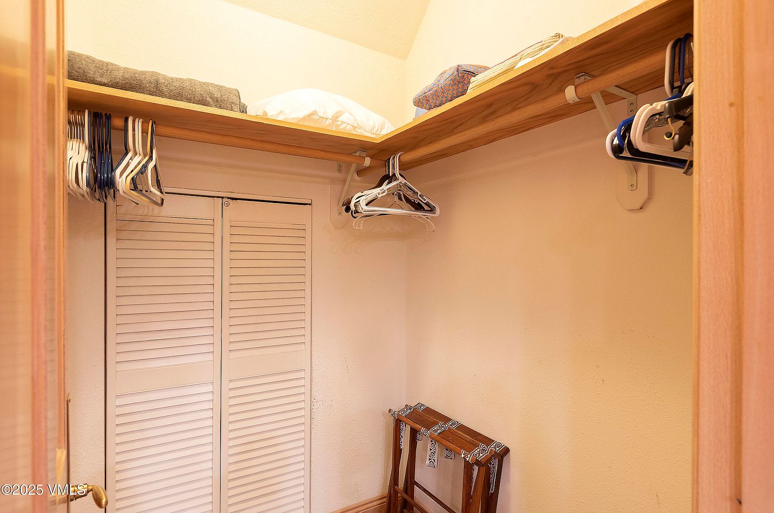 This is an interior shot of a walk-in closet. The closet features a wooden shelf with folded towels and a pillow, along with hanging clothes on hangers. A wooden luggage rack sits on the floor, and the closet has bi-fold doors with horizontal slats.