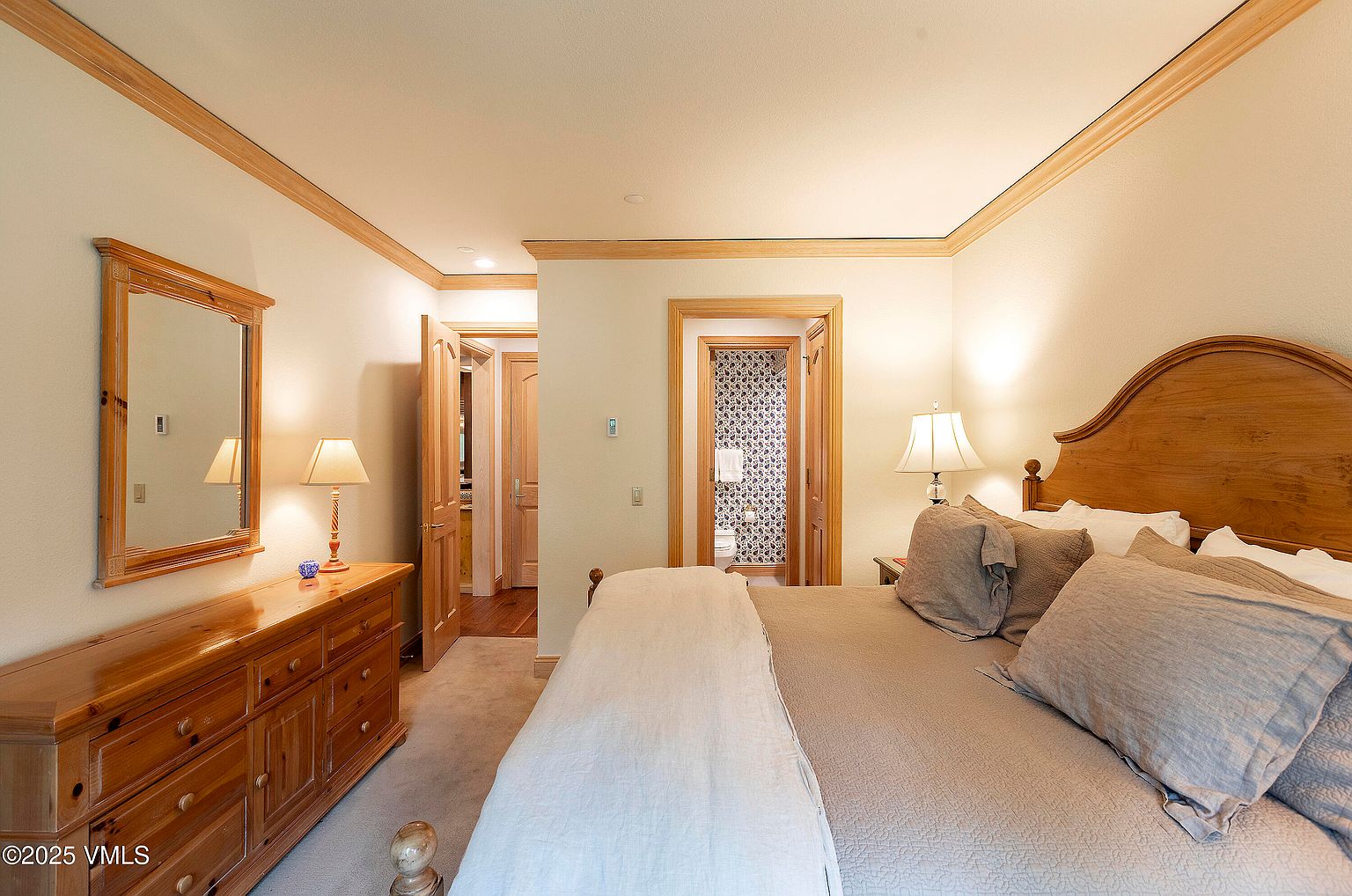 This is a primary bedroom featuring a wooden headboard bed with neutral-toned bedding and pillows. A wooden dresser with a mirror and lamp sits against the wall, and doorways lead to other rooms, including one with patterned wallpaper in the bathroom. The room has a warm and inviting feel with natural wood accents.