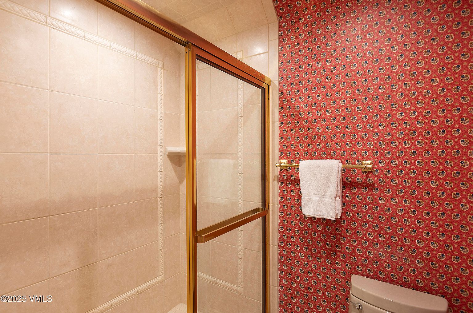 This bathroom features a shower with a gold-framed glass door and beige tile walls. The wallpaper is red with a floral pattern, and a gold towel rack holds a white towel. The overall impression is dated but clean.