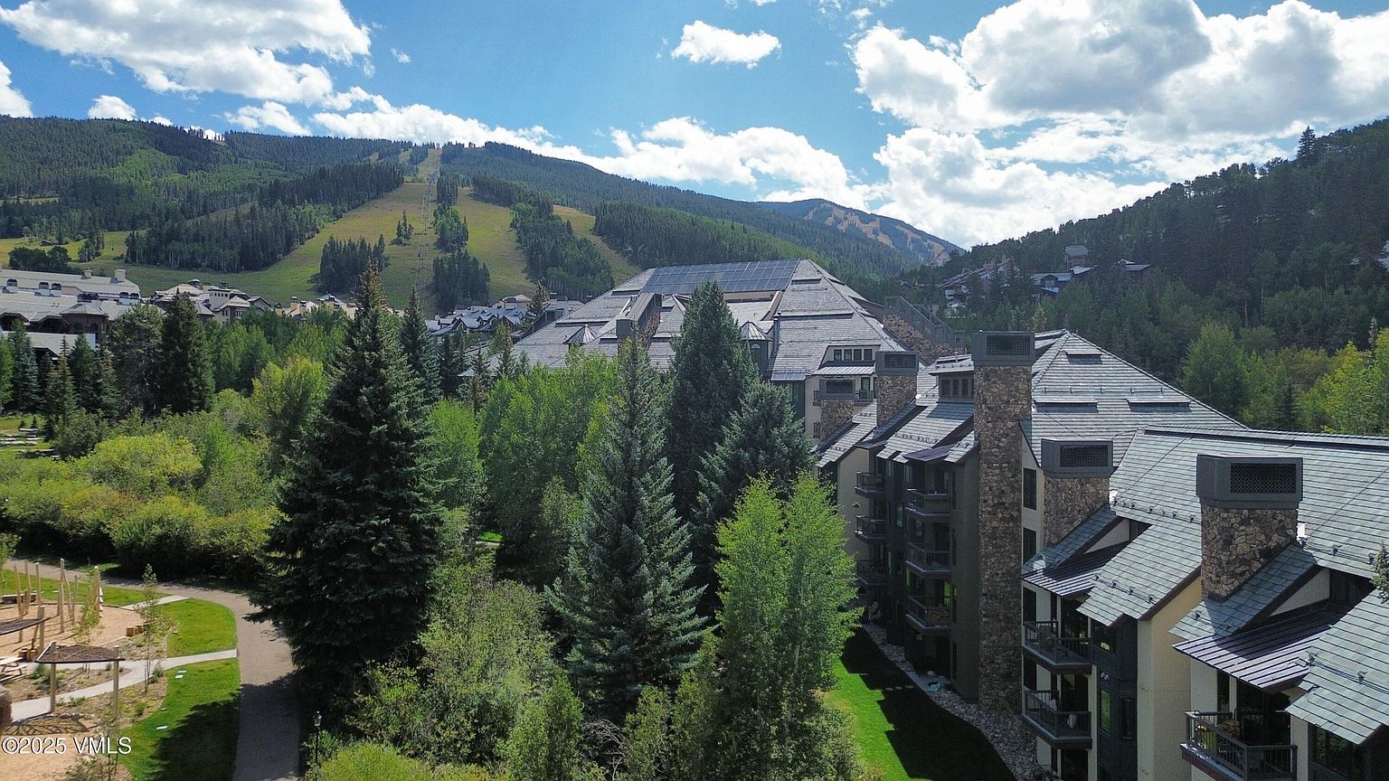 This aerial view showcases a luxurious mountain resort nestled among lush green trees and rolling hills. The architecture features a mix of stone and dark roofing, blending seamlessly with the natural surroundings. A ski slope is visible in the background, suggesting a prime location for winter sports enthusiasts.