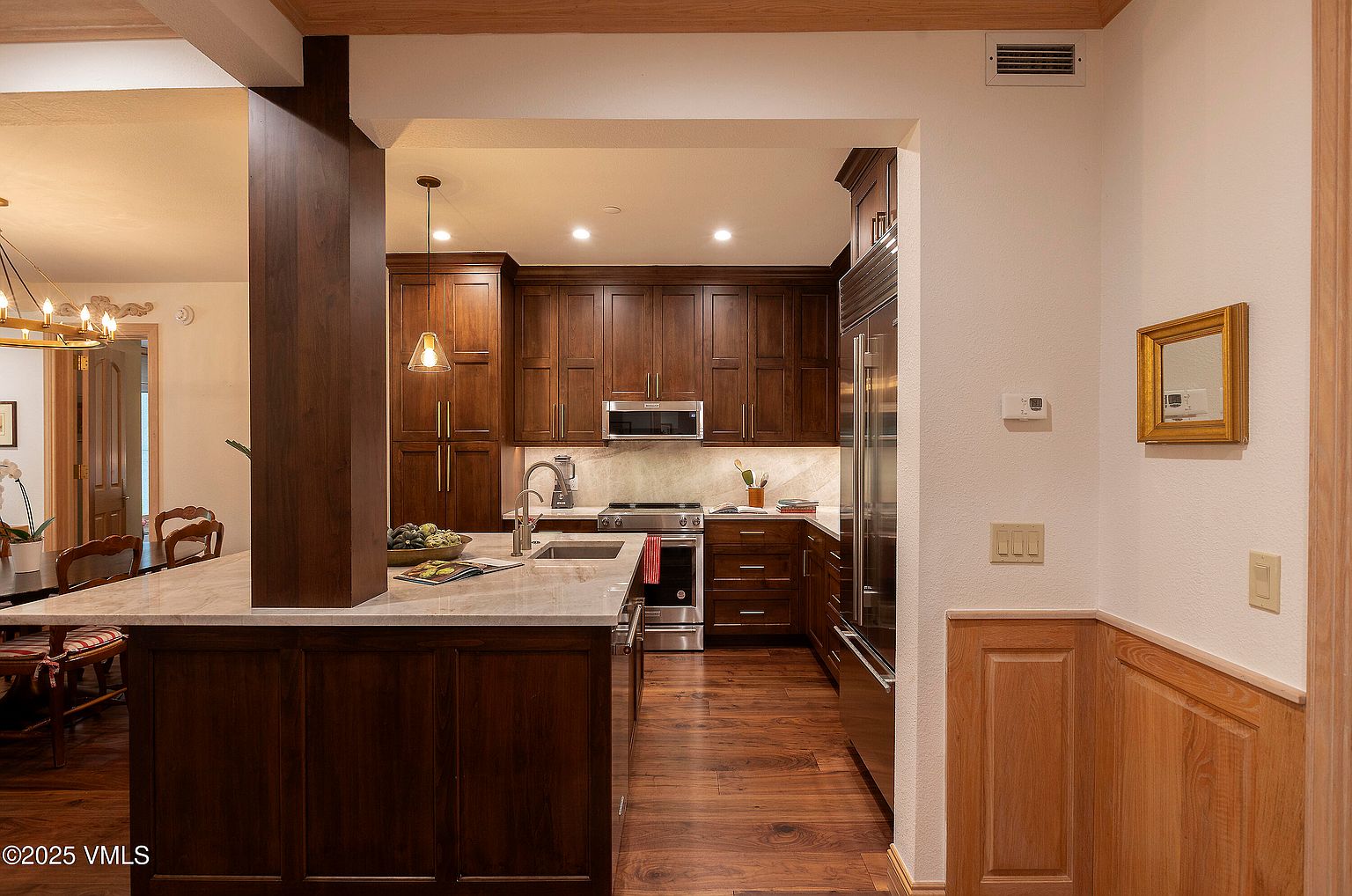 This is a well-appointed kitchen featuring dark wood cabinetry, stainless steel appliances, and light-colored countertops. The kitchen is open to a dining area, creating a seamless flow. The warm wood tones and ample lighting create an inviting and sophisticated atmosphere.