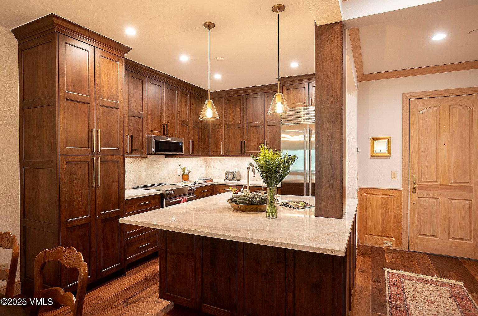 This is a warm and inviting kitchen featuring rich, dark wood cabinetry and a light-colored marble countertop island. Stainless steel appliances are integrated seamlessly into the design, and pendant lights hang above the island, providing focused illumination. The kitchen exudes a sense of classic elegance and functionality.