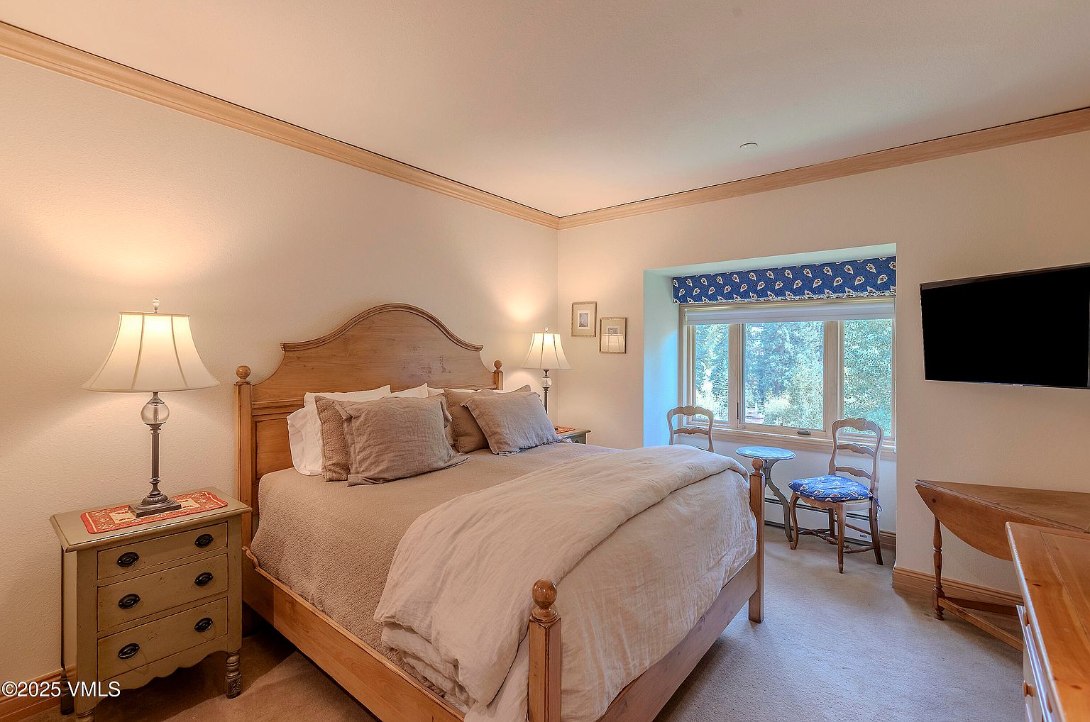 This is a cozy primary bedroom featuring a wooden bed frame with neutral-toned bedding and pillows. A bedside table with a lamp sits to the left, while a window with a blue valance and two chairs are visible in the background. A wooden desk is on the right side of the room. The room has a warm and inviting atmosphere.