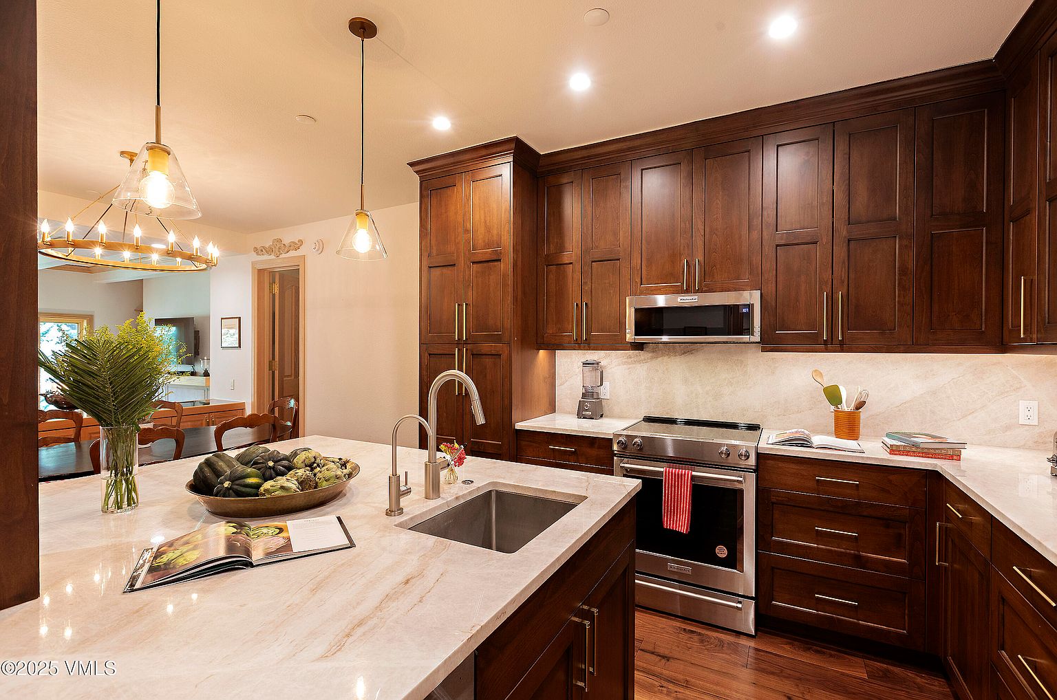 This is a well-appointed kitchen featuring dark wood cabinetry, stainless steel appliances, and a marble countertop island with a sink. The kitchen has a warm and inviting feel, with pendant lighting and a view into the dining area. The hardwood floors add to the overall elegance of the space.