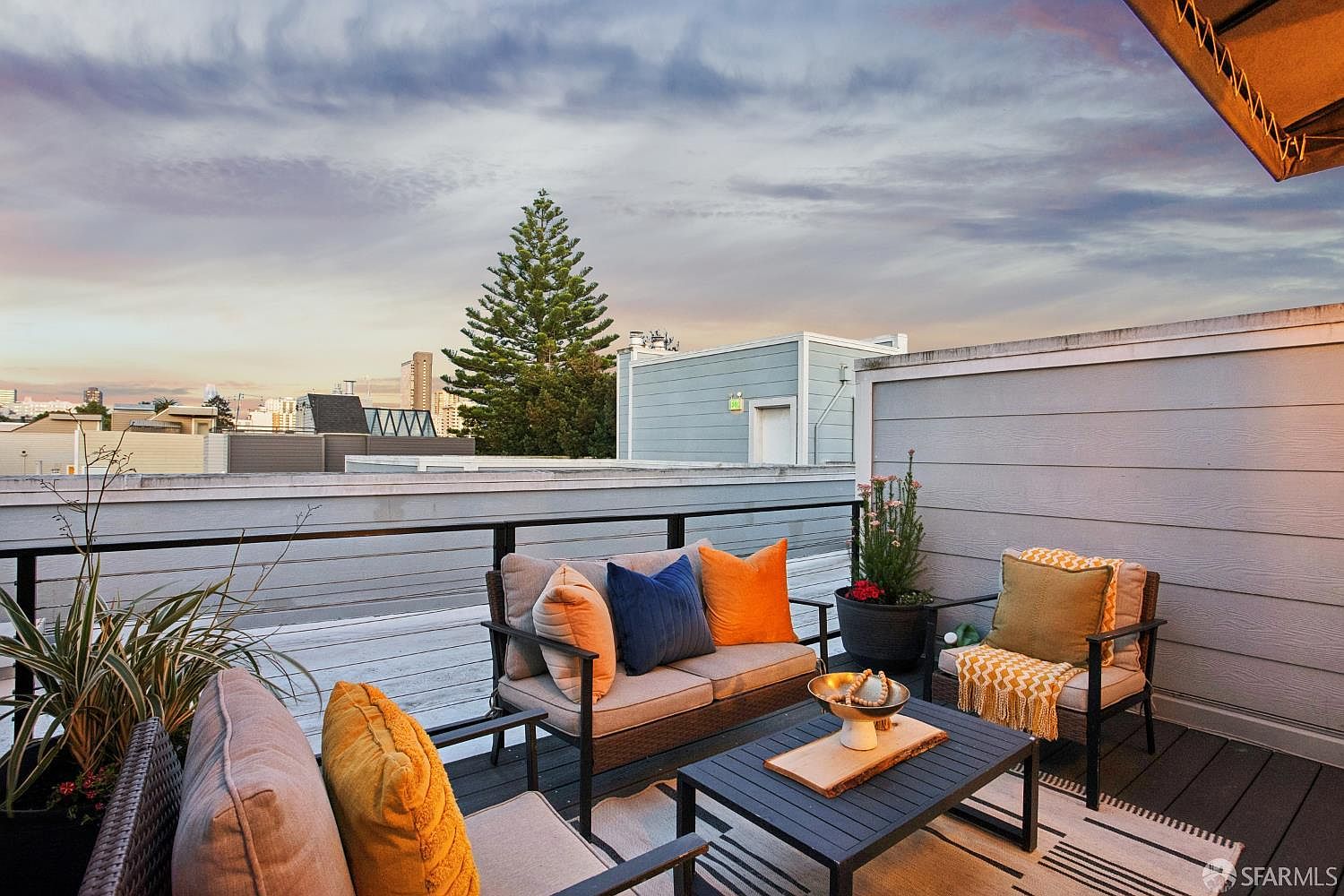 This inviting rooftop deck features a comfortable outdoor seating area with a sofa and armchair arranged around a modern coffee table. The space is decorated with vibrant orange and blue accent pillows, potted plants, and a patterned area rug, creating a cozy atmosphere against a backdrop of city skyline views. The perspective is a wide-angle shot from a slightly elevated position, capturing the relaxing urban outdoor living space at dusk.