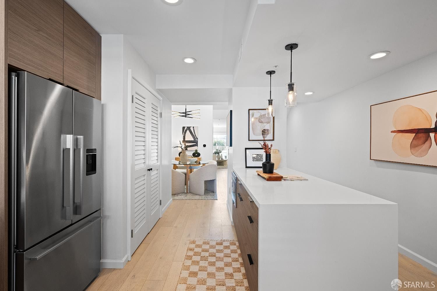 This modern kitchen features a sleek island with a white countertop and wood-toned cabinetry, complemented by stainless steel appliances and minimalist pendant lighting. The perspective looks down a bright, open hallway toward a dining area, creating a seamless flow between spaces. The light wood flooring and neutral color palette contribute to a clean, contemporary, and inviting atmosphere.