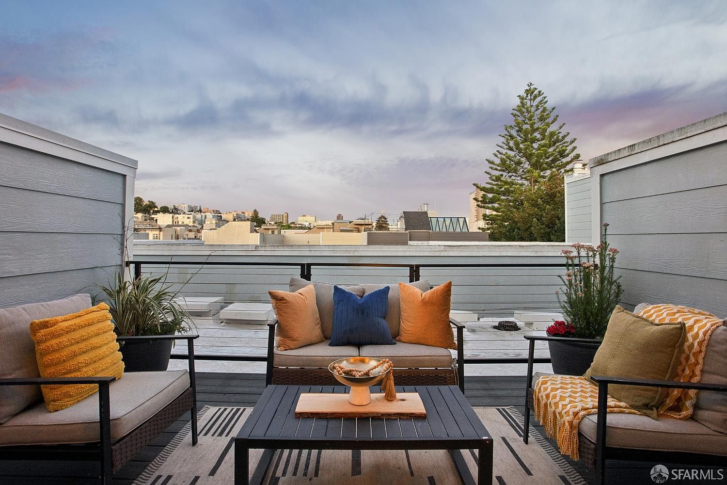 This inviting rooftop deck features a comfortable seating arrangement with a loveseat and two armchairs centered around a modern coffee table. The space is decorated with plush cushions, a patterned area rug, and potted plants, creating a cozy outdoor living area. The deck offers a panoramic view of the surrounding city skyline under a dramatic, cloud-filled sky, making it an ideal spot for relaxation or entertaining.