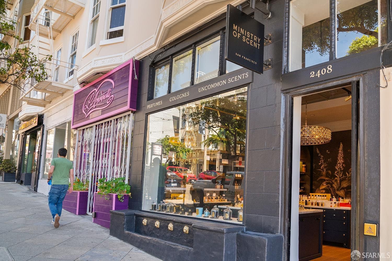 This street-level view captures the storefront of 'Ministry of Scent' in San Francisco, featuring a dark, modern facade with large display windows showcasing perfume bottles. A person walks along the sidewalk past the shop, which is situated next to a vibrant purple storefront, creating a lively urban retail atmosphere. The perspective is a wide-angle shot that highlights the architectural details of the building's exterior and the inviting, well-lit interior visible through the open doorway.