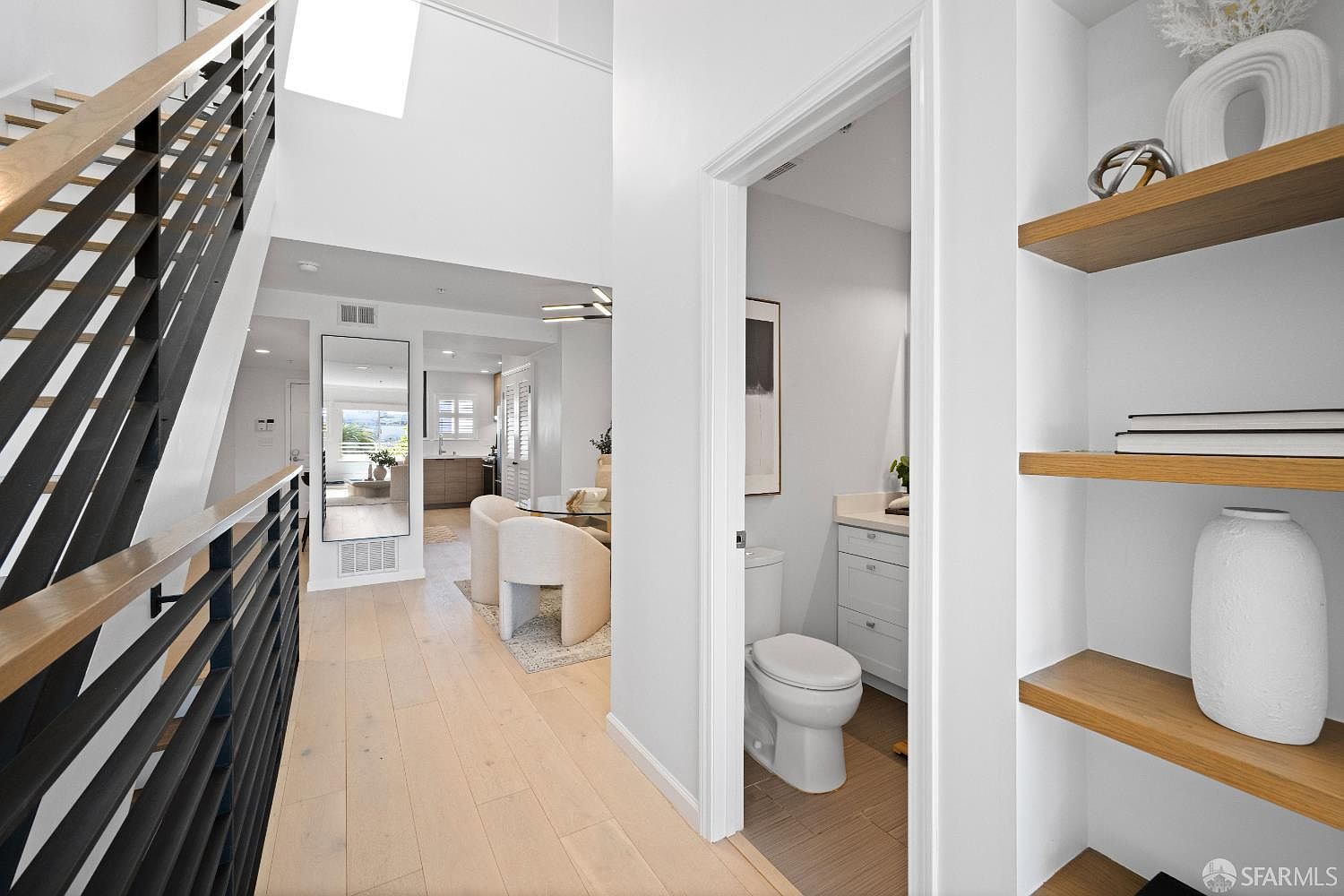 This bright, modern hallway features light hardwood flooring and a striking staircase with black metal railings and wooden handrails. To the right, built-in wooden shelves display decorative items, while a doorway leads into a clean, white-tiled bathroom. The perspective looks through the hallway toward a dining area, creating an open and airy atmosphere with a contemporary aesthetic.
