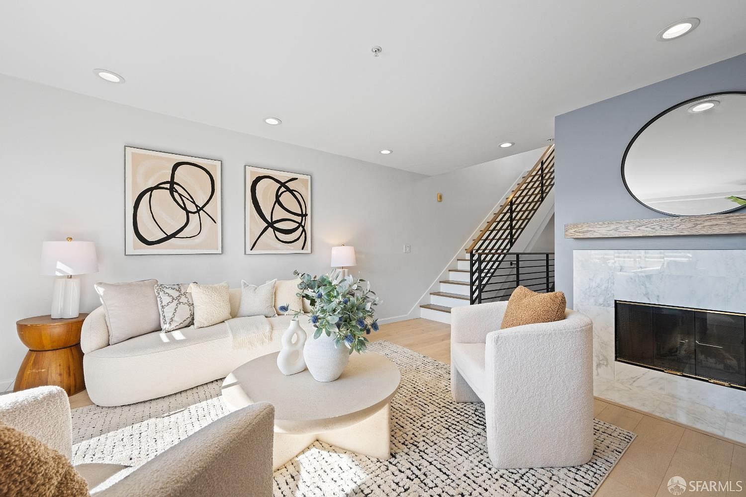 This bright and airy living room features a contemporary design with a neutral color palette, highlighted by a plush cream-colored sofa and matching armchairs. A marble-faced fireplace serves as a focal point, complemented by a large circular mirror and abstract wall art, while a staircase with modern railings adds architectural interest in the background. The space is bathed in natural light, creating a warm and inviting atmosphere perfect for relaxation.