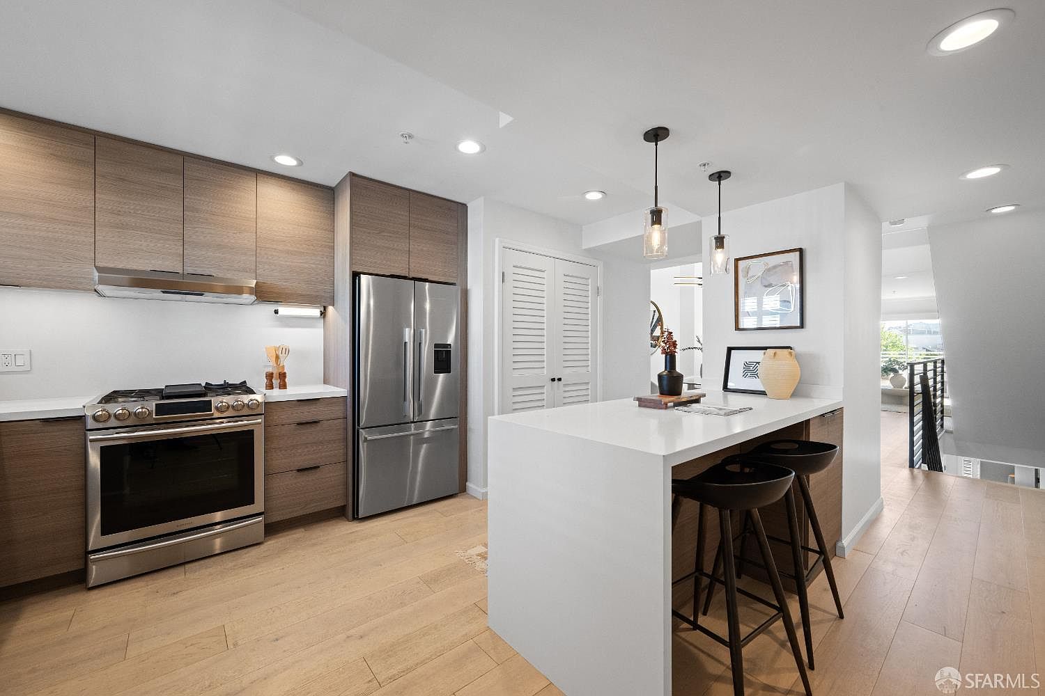 This modern, open-concept kitchen features sleek wood-toned cabinetry, stainless steel appliances, and a large white quartz waterfall island with bar seating. The space is illuminated by stylish pendant lights and recessed lighting, creating a bright and inviting atmosphere. Light-colored hardwood flooring flows throughout, leading toward a staircase in the background, emphasizing a contemporary and functional design.