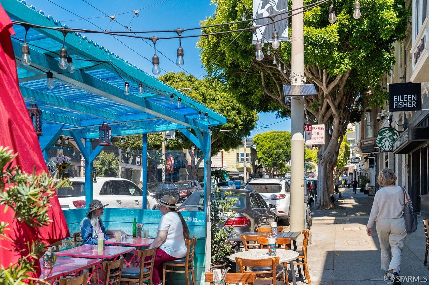 This vibrant outdoor dining scene captures a lively street-side patio area featuring bright blue wooden structures and string lights. The perspective is from the sidewalk, showcasing patrons enjoying a meal while pedestrians walk by, with local storefronts like Eileen Fisher and Starbucks visible in the background. The atmosphere is bustling and urban, highlighting the property's prime location in a walkable, commercial neighborhood.
