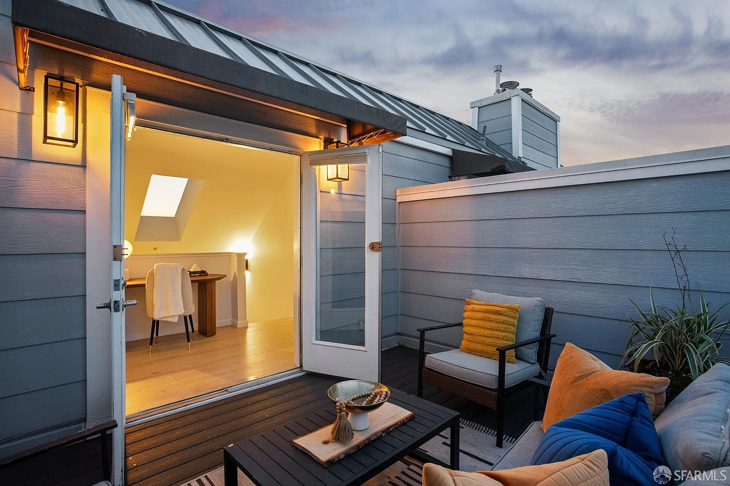 This inviting rooftop deck features a modern, cozy seating area with a dark wood coffee table, a comfortable armchair with a yellow accent pillow, and a plush sofa with colorful cushions. The deck seamlessly connects to a bright, minimalist home office space through open French doors, creating an ideal indoor-outdoor flow. The scene is captured at dusk, with warm exterior lighting and a soft, twilight sky providing a serene and sophisticated atmosphere.
