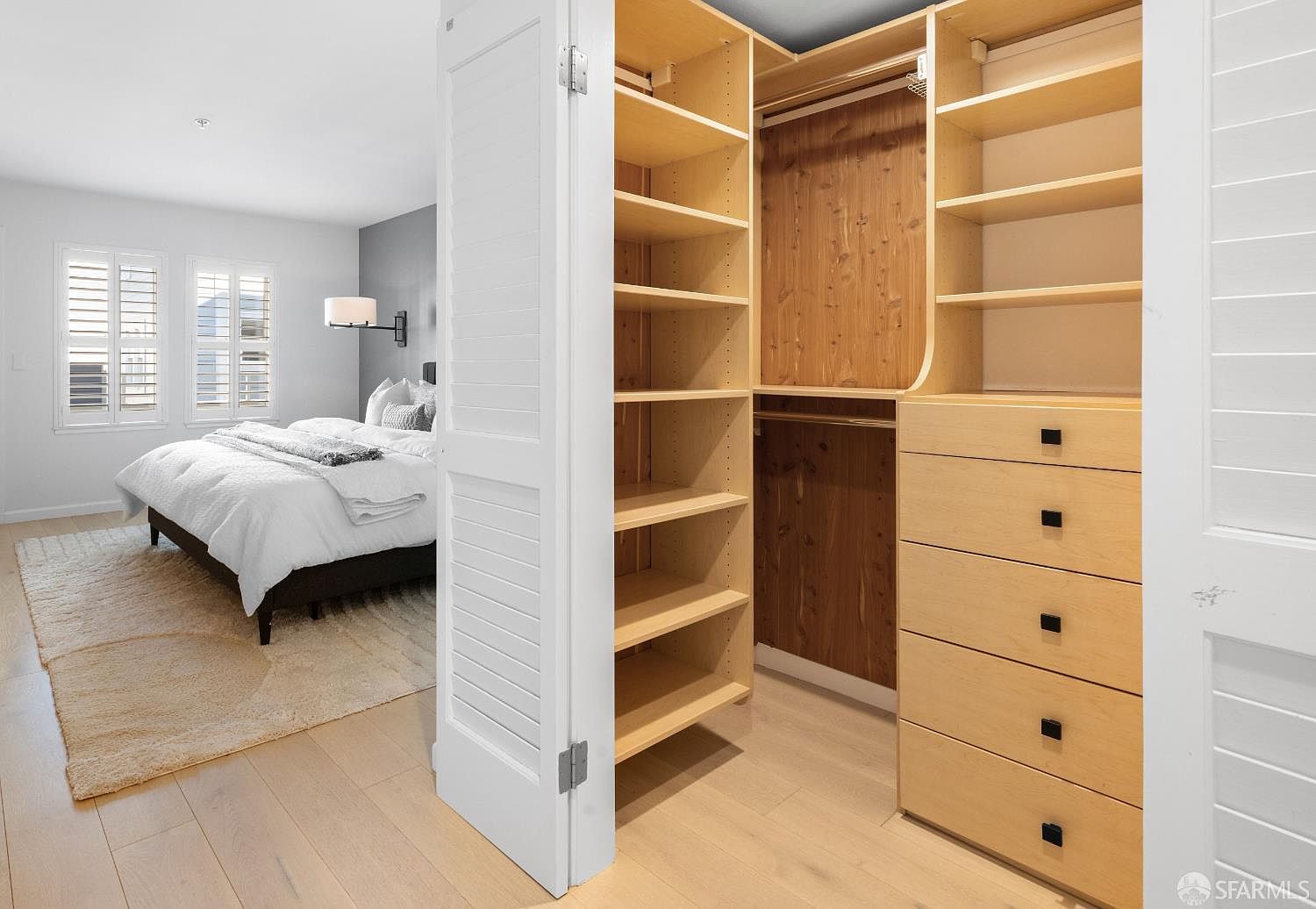 This image captures a view from a bright, modern bedroom looking into a spacious walk-in closet. The closet features custom light-wood shelving, hanging rods, and a bank of drawers with sleek black hardware, while the bedroom is furnished with a plush bed and neutral-toned decor. The perspective highlights the seamless transition between the sleeping area and the organized storage space, emphasizing a clean and contemporary aesthetic.