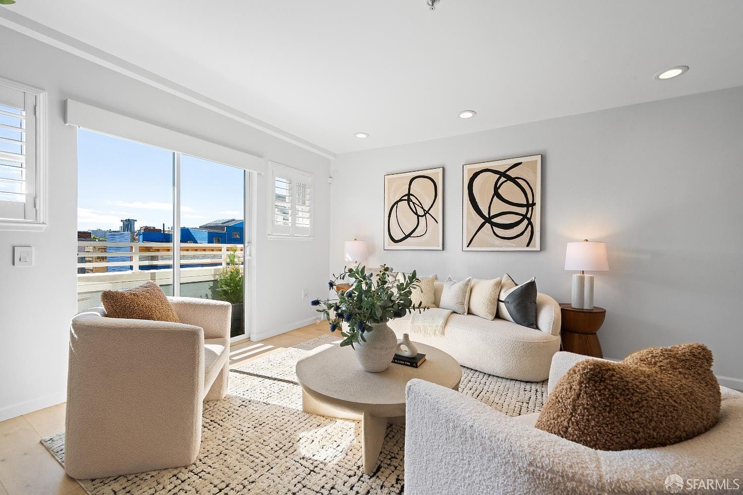 This bright and airy living room features a contemporary, neutral aesthetic with a curved sofa, two plush armchairs, and a textured area rug. A large sliding glass door provides abundant natural light and access to an outdoor balcony, while abstract wall art adds a sophisticated touch to the space. The room is anchored by a central round coffee table, creating a warm and inviting atmosphere perfect for relaxation.