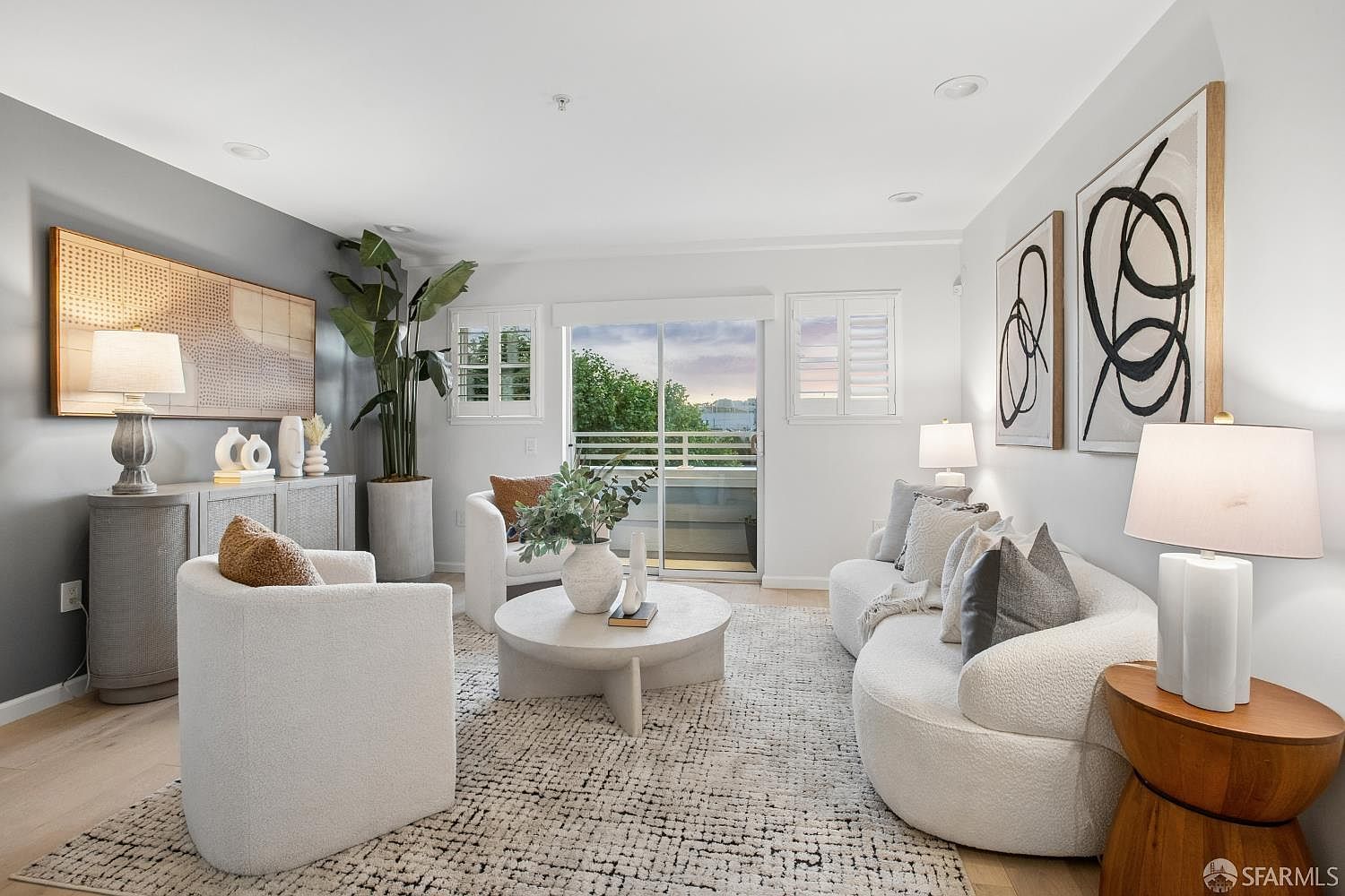 This bright and airy living room features a contemporary design with a curved white sofa, matching armchairs, and a textured area rug. The space is accented by a large potted plant, abstract wall art, and a sleek round coffee table, all bathed in natural light from the sliding glass door leading to a balcony. The neutral color palette and sophisticated furniture arrangement create a welcoming and upscale atmosphere.