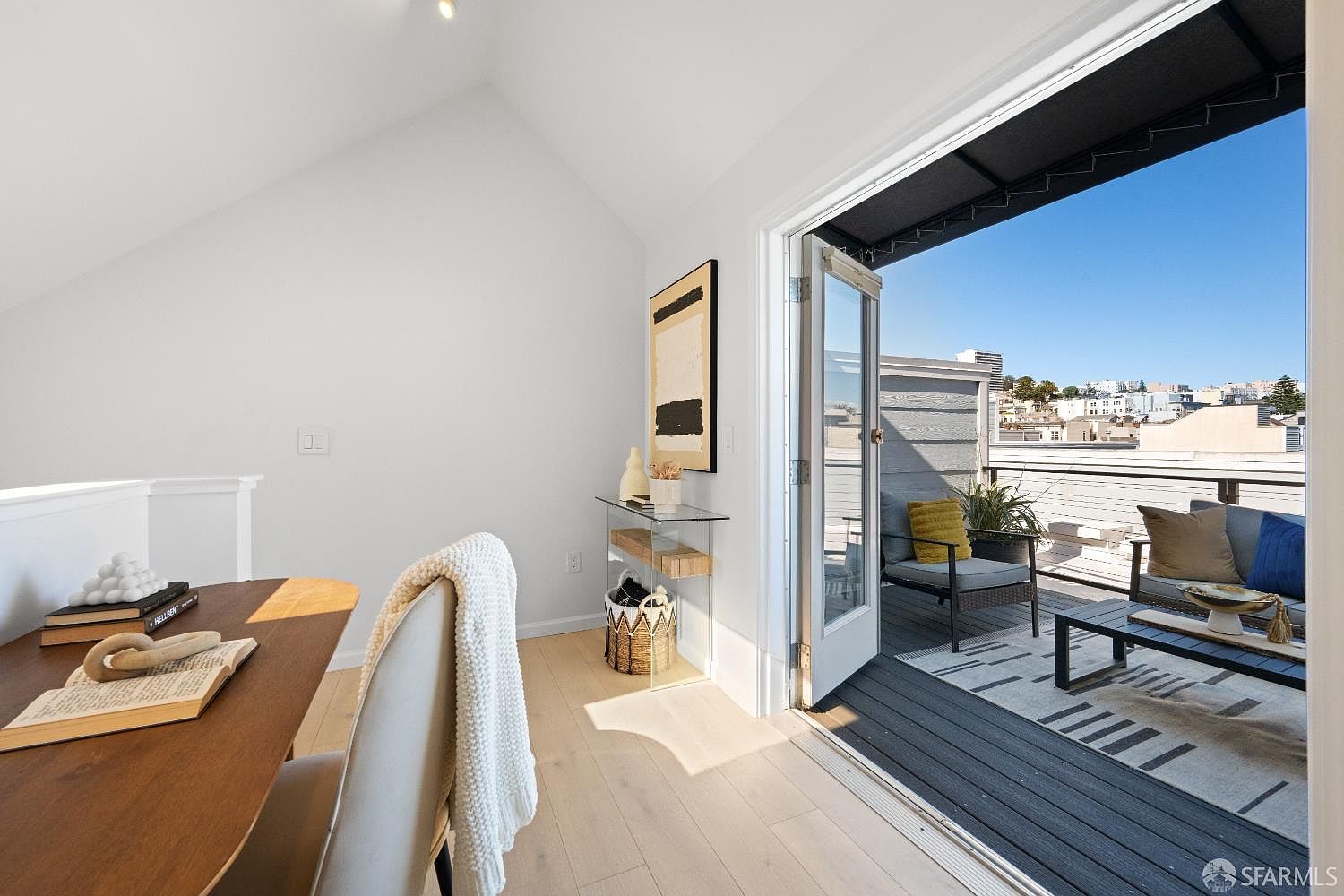 This bright and airy home office space features a sleek wooden desk in the foreground and opens directly onto a private outdoor deck through glass doors. The room is decorated with minimalist, modern accents, including a glass console table and abstract wall art, creating a seamless transition between indoor and outdoor living. The light wood flooring and vaulted ceilings enhance the sense of openness, making it an inviting and productive environment.