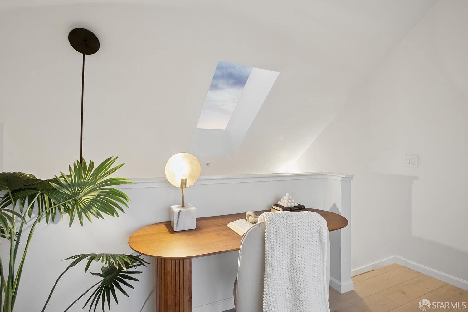 This cozy and modern home office nook is nestled under a sloped ceiling with a skylight that floods the space with natural light. The area features a sleek, oval-shaped wooden desk, a contemporary lamp with a marble base, and a comfortable chair draped with a soft white throw blanket. A lush potted plant adds a touch of greenery, creating a serene and productive atmosphere.