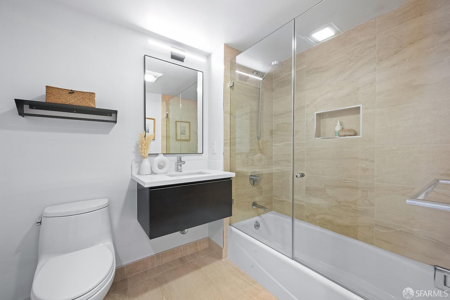 This modern, compact bathroom features a sleek floating vanity with a white countertop and a contemporary mirror. The shower-tub combination is enclosed by a frameless glass door and finished with neutral-toned, large-format wall tiles. The space is bright and clean, utilizing a minimalist aesthetic that maximizes the functionality of the room.