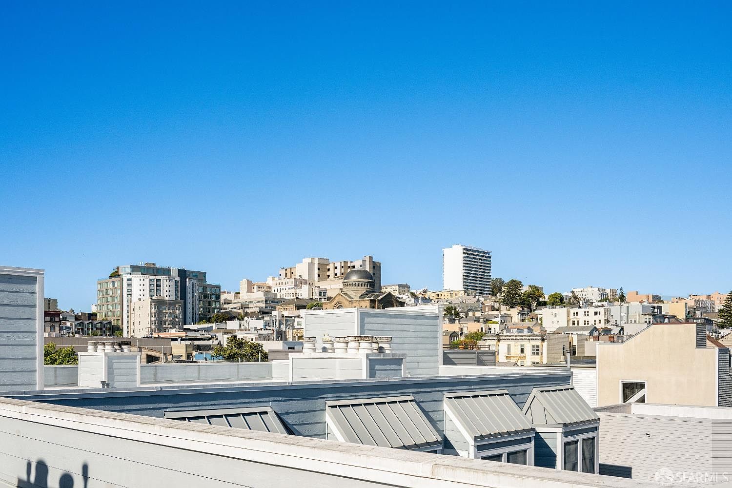 This image captures a wide-angle view from a rooftop deck, showcasing a panoramic cityscape under a clear blue sky. The foreground features modern architectural roof elements with metal awnings, while the background reveals a dense urban environment with various residential and commercial buildings. The perspective provides a sense of height and openness, highlighting the property's elevated outdoor living space.