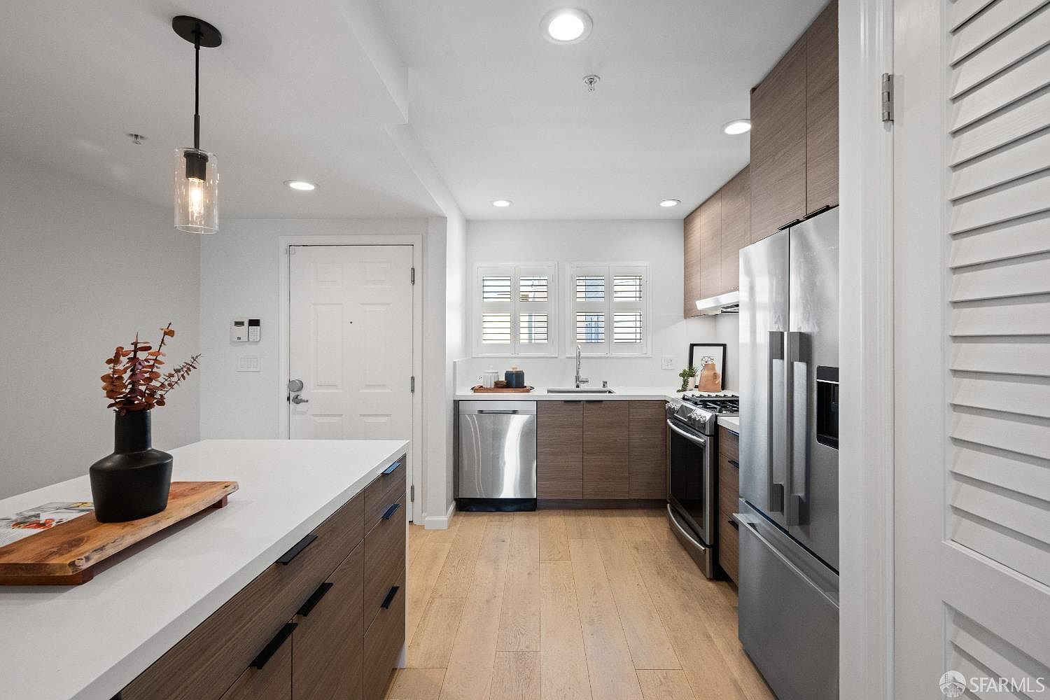 This modern kitchen features sleek, wood-toned cabinetry paired with white quartz countertops and stainless steel appliances. A central island with a decorative vase and wooden serving board serves as a focal point, while a pendant light adds a touch of warmth to the space. The open-concept design flows seamlessly into the entryway, creating a bright and inviting atmosphere with light wood flooring throughout.