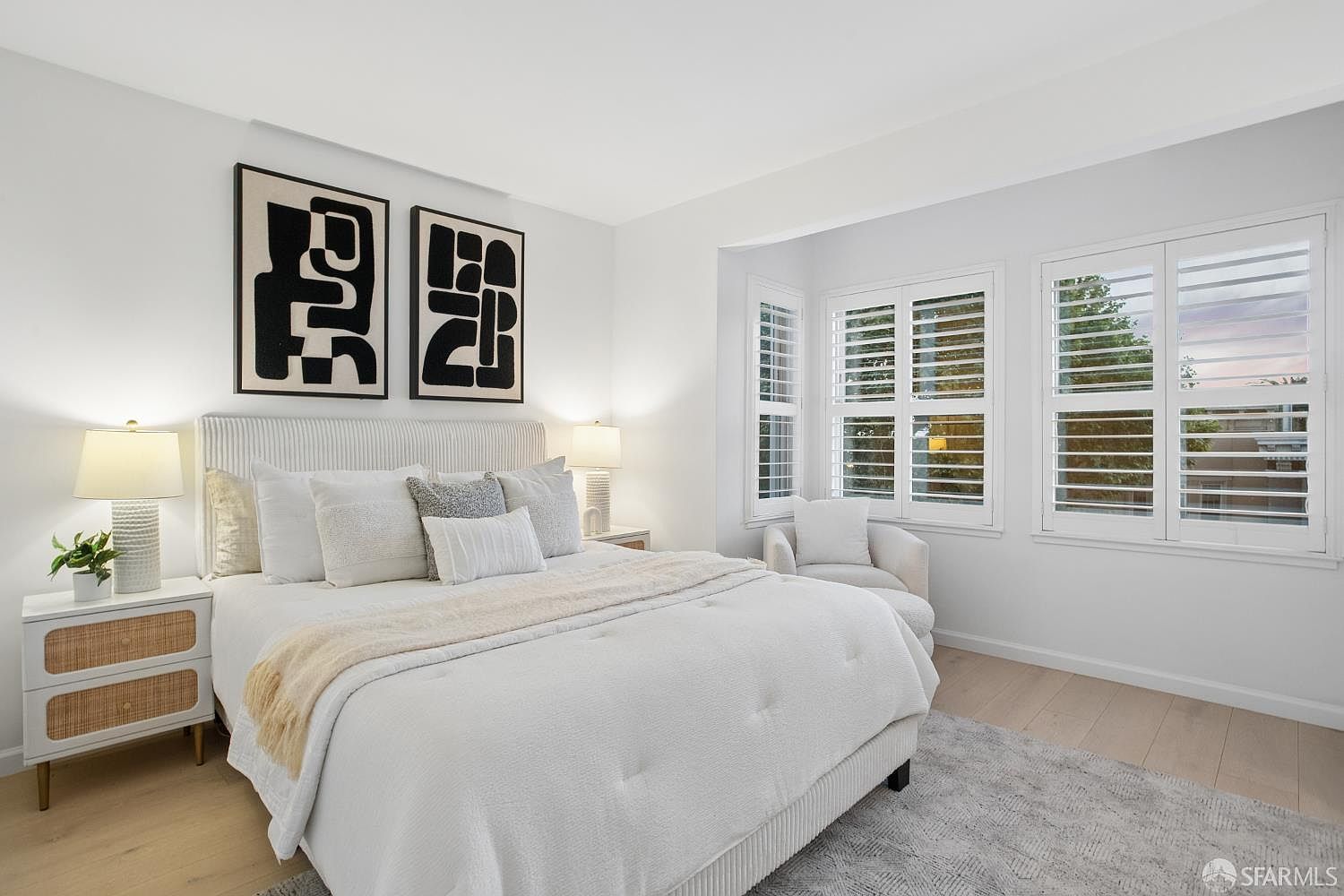 This bright and airy primary bedroom features a neutral color palette with a plush upholstered bed as the focal point, complemented by modern abstract wall art. The room includes a cozy reading nook by the window with plantation shutters and light wood flooring, creating a serene and inviting atmosphere. The perspective is a straight-on shot that captures the symmetry and clean, contemporary design of the space.