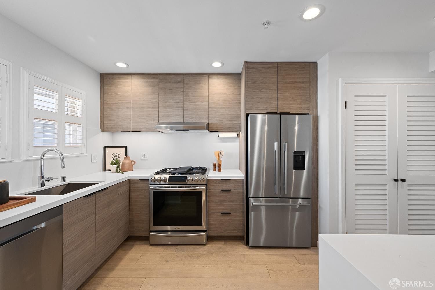 This modern, U-shaped kitchen features sleek wood-grain cabinetry paired with clean white countertops and a matching backsplash. The space is equipped with high-end stainless steel appliances, including a French-door refrigerator and a gas range, all illuminated by recessed ceiling lights and under-cabinet lighting. The light wood-toned flooring and minimalist design create a bright, contemporary, and functional culinary environment.