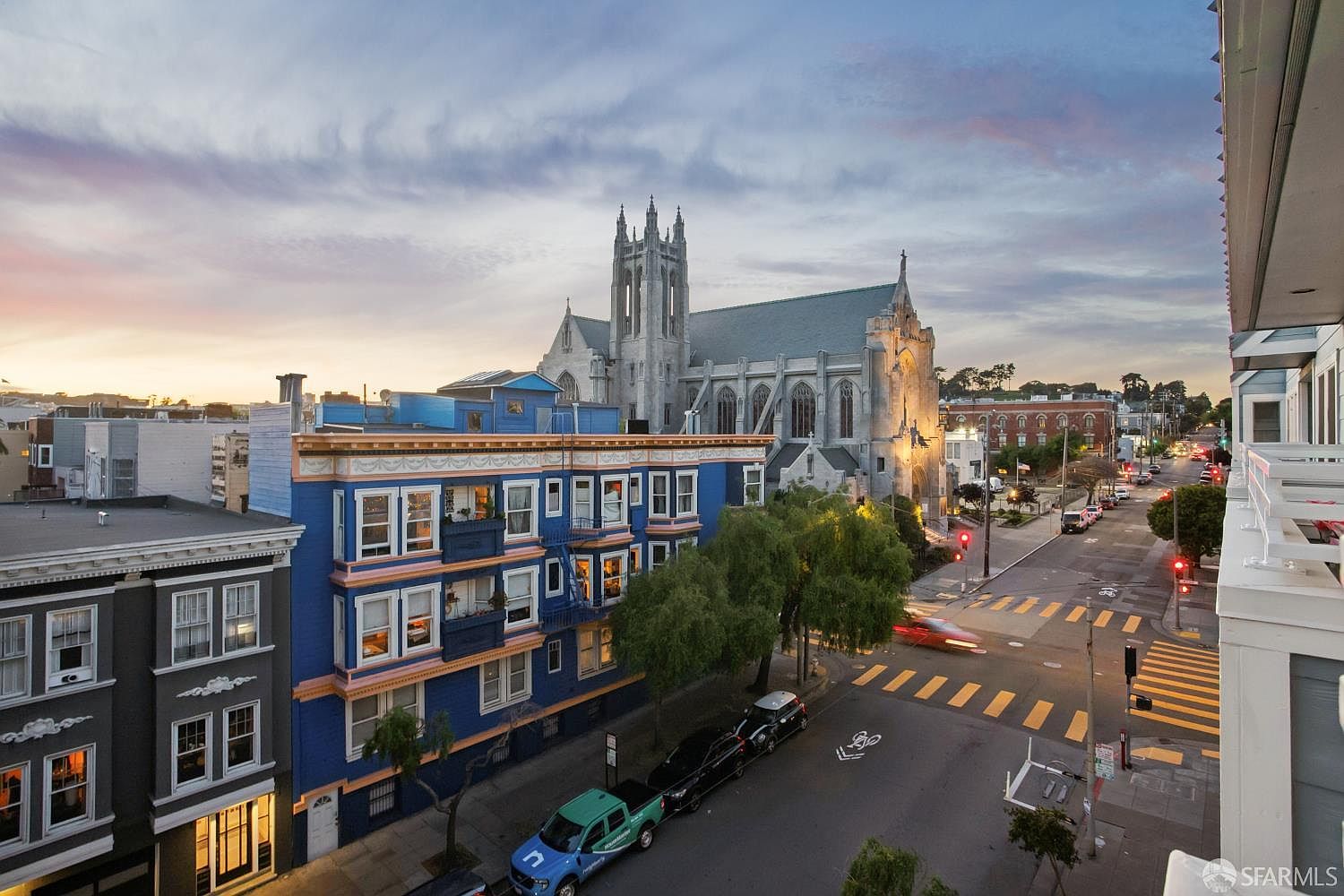 This elevated exterior view captures a vibrant urban streetscape in San Francisco, featuring a striking blue multi-story residential building in the foreground. A prominent Gothic-style stone church stands majestically in the background, set against a soft, golden-hour sunset sky. The perspective provides a cinematic overview of the neighborhood, highlighting the intersection, street traffic, and the architectural contrast between the historic church and the colorful city homes.