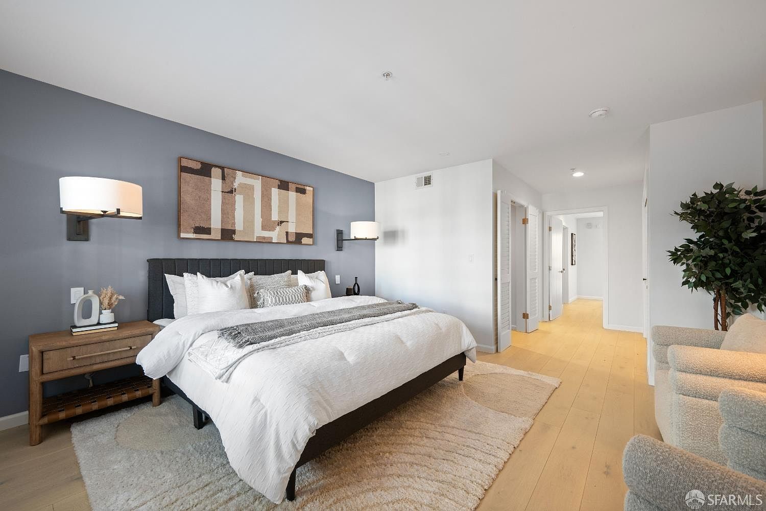 This spacious primary bedroom features a modern aesthetic with a large bed centered against a striking blue accent wall adorned with abstract art. The room is bright and airy, highlighted by light wood flooring, a textured area rug, and two comfortable armchairs in the foreground. The perspective captures the inviting layout, leading the eye toward the hallway and open doorways, creating a sense of flow and comfort.
