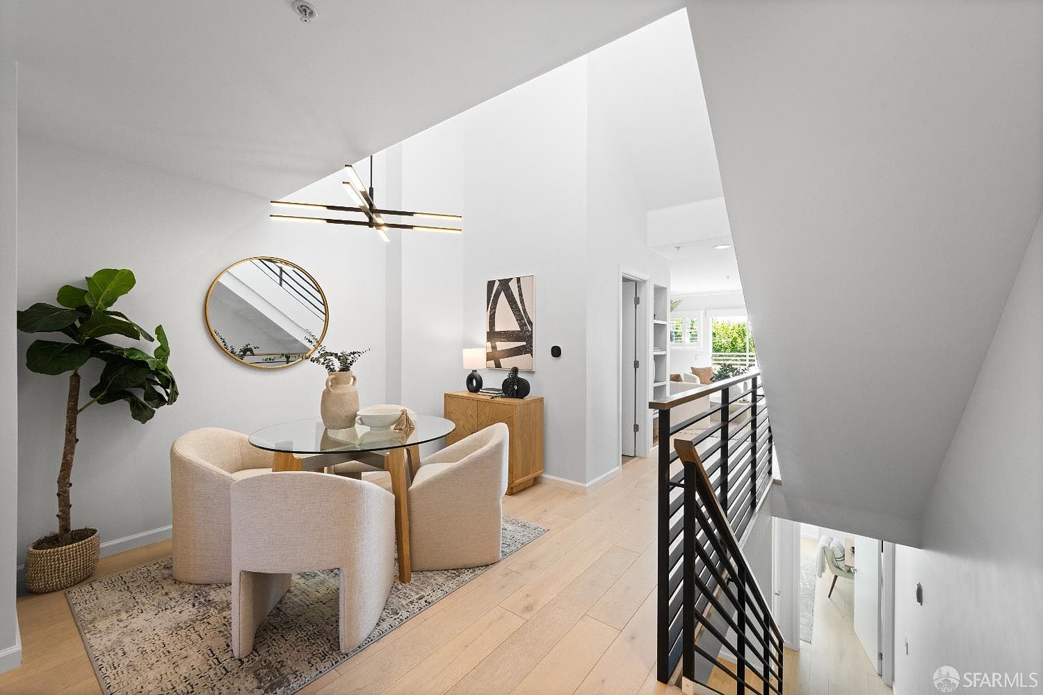 This bright and airy dining area features a modern glass-topped table surrounded by three cream-colored upholstered chairs, set atop a textured area rug. A striking contemporary chandelier hangs above, while a round mirror and a sleek wooden sideboard add sophisticated touches to the space. The perspective looks down from a staircase landing, highlighting the open-concept layout and the seamless transition to the living area beyond.