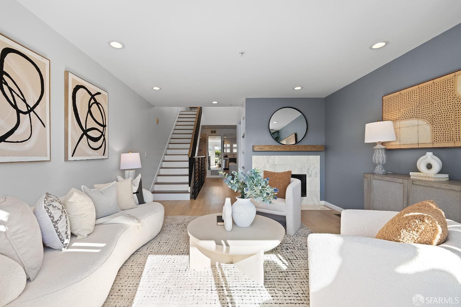 This bright and airy living room features a contemporary design with a neutral color palette, highlighted by a curved sofa and a marble-faced fireplace. The space is accented by modern abstract wall art, a round coffee table, and a staircase leading to the upper level, creating an inviting and sophisticated atmosphere. The perspective is a wide-angle shot from the center of the room, emphasizing the open-concept layout and elegant decor.