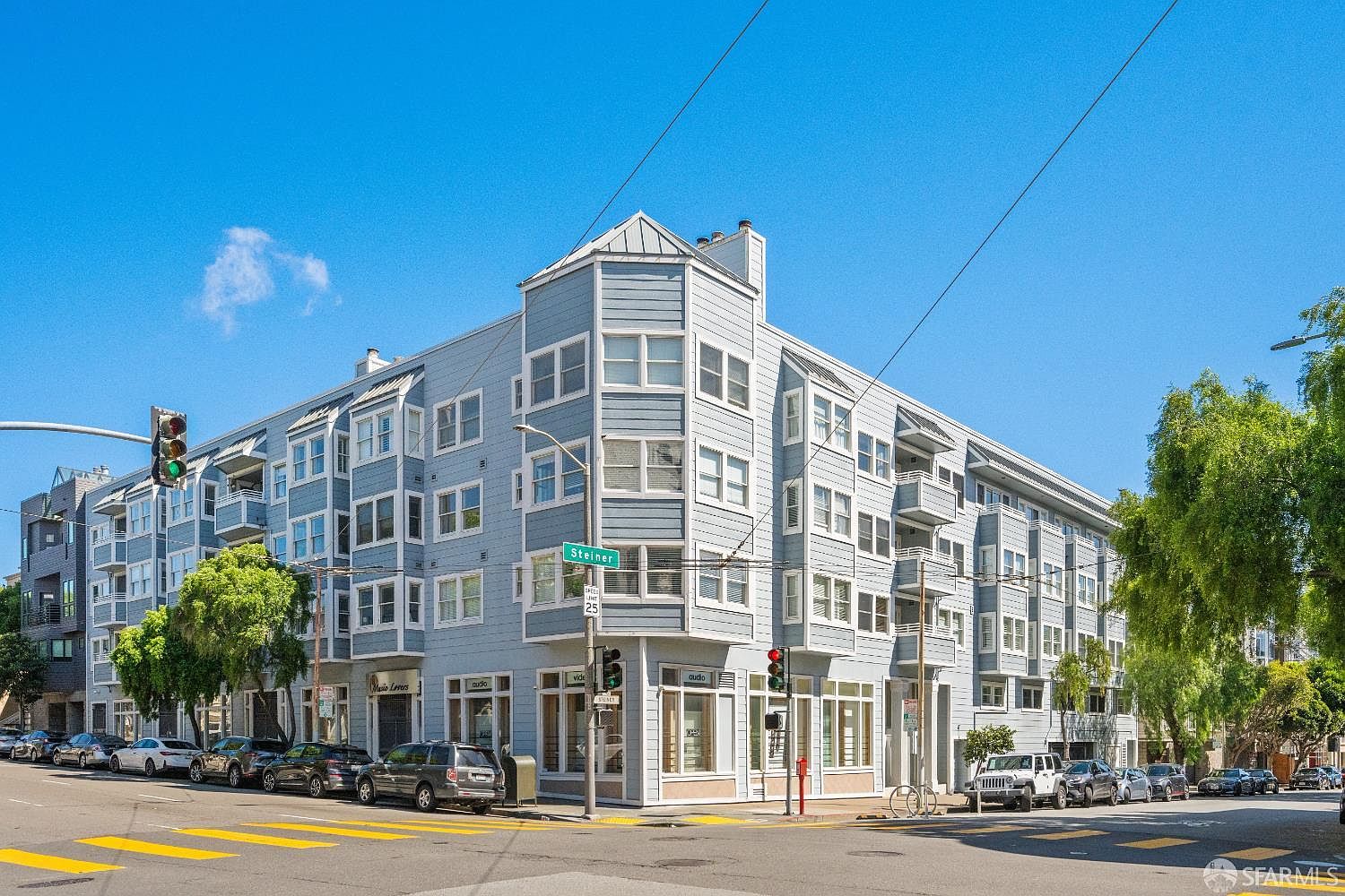 This image captures a prominent corner-lot multi-story residential building in an urban setting, featuring light blue siding and a classic architectural design with bay windows. The perspective is a wide-angle street-level shot, showcasing the building's integration into the neighborhood with storefronts on the ground level and residential units above. The clear blue sky and bright daylight emphasize the building's clean lines and well-maintained facade.
