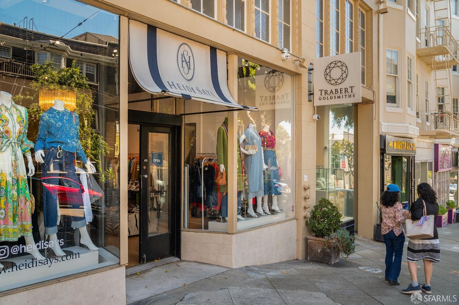 This street-level view captures the storefronts of 'Heidi Says' and 'Trabert Goldsmiths' along a bustling urban sidewalk. The scene features large display windows showcasing mannequins in fashionable attire, with a classic striped awning over the entrance and a prominent sign for the jewelry store. Two pedestrians are walking past the shops, adding a sense of local activity and neighborhood charm to the commercial facade.