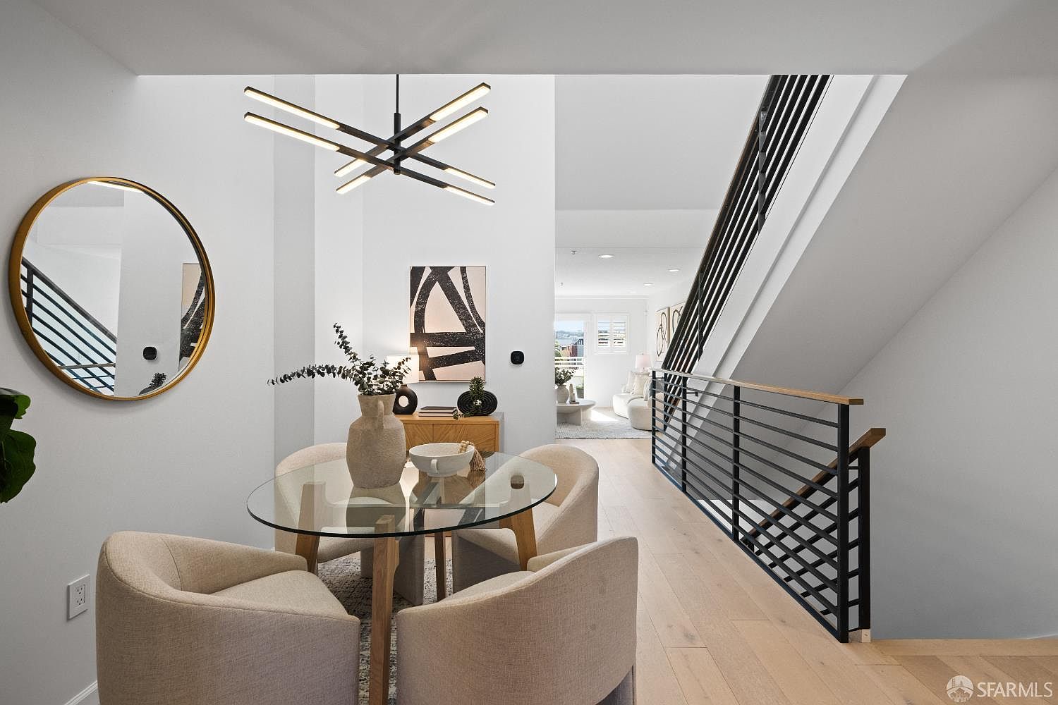 This bright and modern dining area features a glass-topped table surrounded by four upholstered cream chairs, set beneath a striking contemporary geometric chandelier. The space is accented by a large circular gold-framed mirror and abstract wall art, creating a sophisticated and airy atmosphere. A sleek black metal staircase railing leads the eye toward an open-concept living space in the background, emphasizing the home's open floor plan and high-end design.