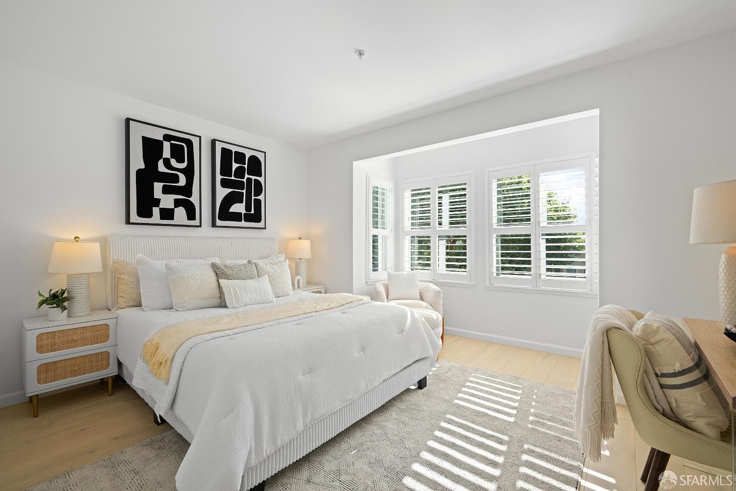 This bright and airy primary bedroom features a neutral color palette with a plush white bed as the focal point, accented by modern abstract art and textured throw pillows. A cozy reading nook is positioned by the large bay window, which is fitted with classic white plantation shutters that allow soft, natural light to fill the room. The space is completed by light wood flooring and a stylish desk area, creating a serene and sophisticated atmosphere.