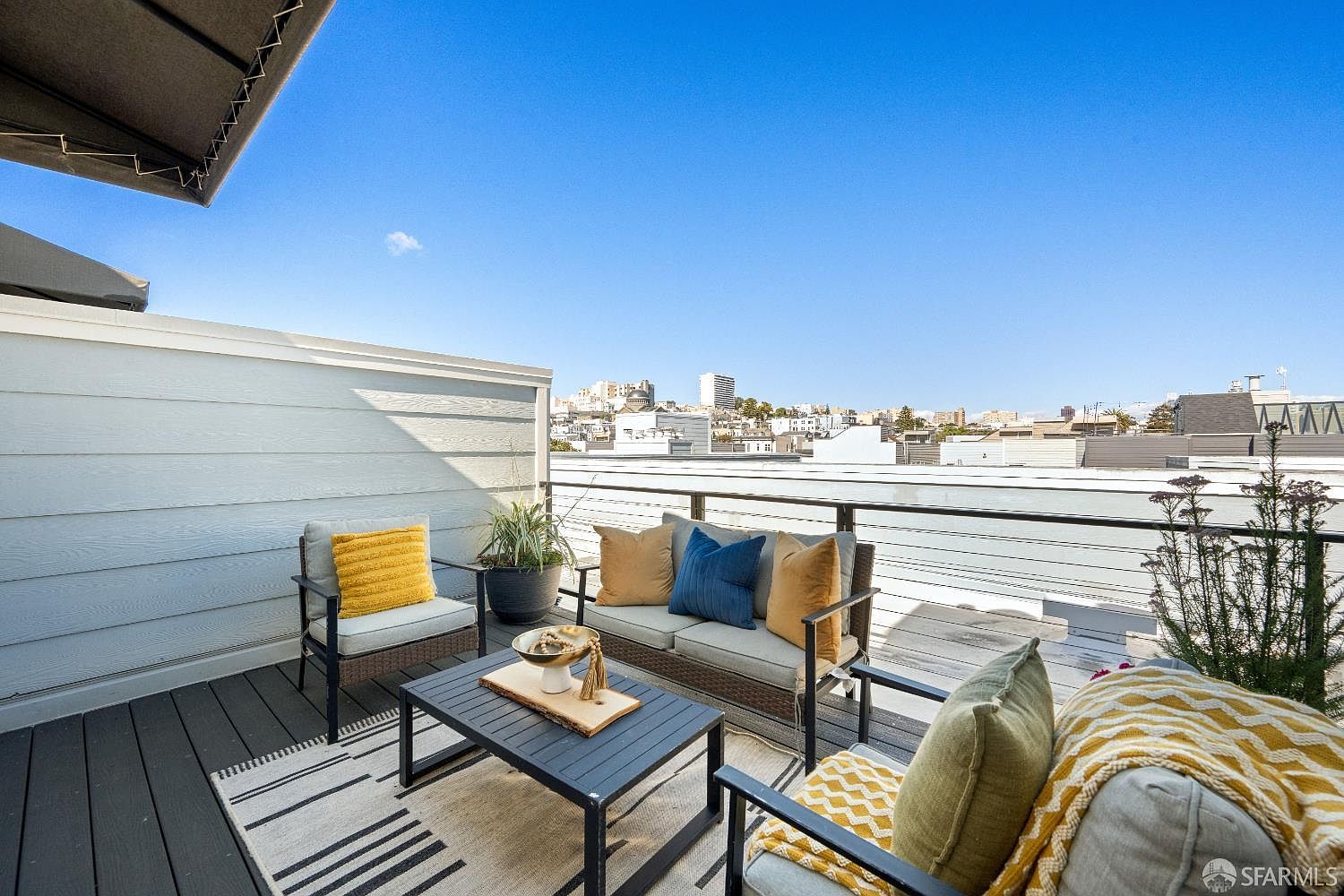 This inviting rooftop deck features a modern outdoor seating arrangement with a sofa, armchair, and coffee table set atop a striped area rug. The space is framed by a sleek metal railing that offers an expansive, unobstructed view of the surrounding city skyline under a clear blue sky. The neutral color palette of the furniture, accented by vibrant yellow and blue cushions, creates a comfortable and stylish urban retreat.